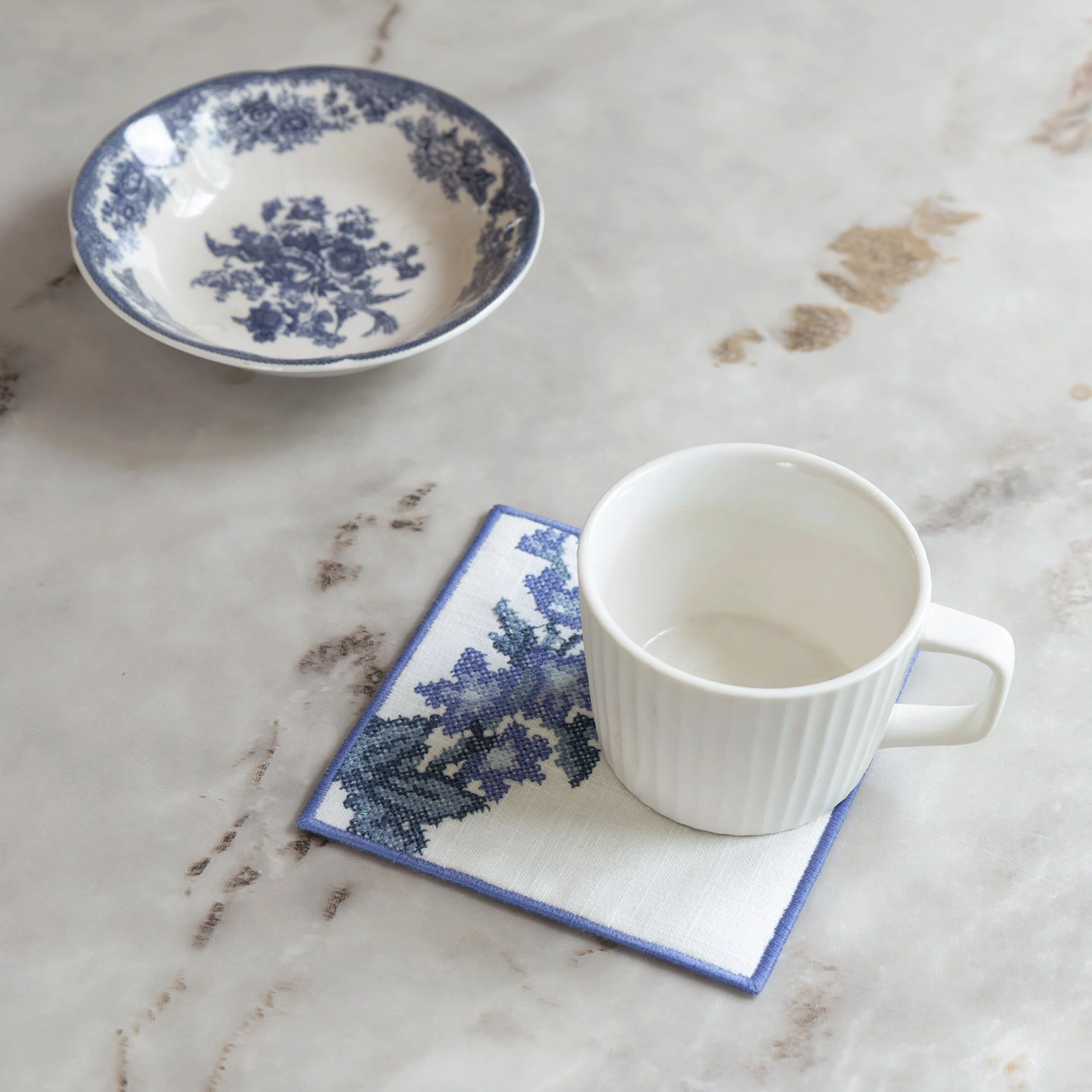 Cross stitch floral coaster with lavender embroidery beneath a white ceramic cup on a marble tabletop