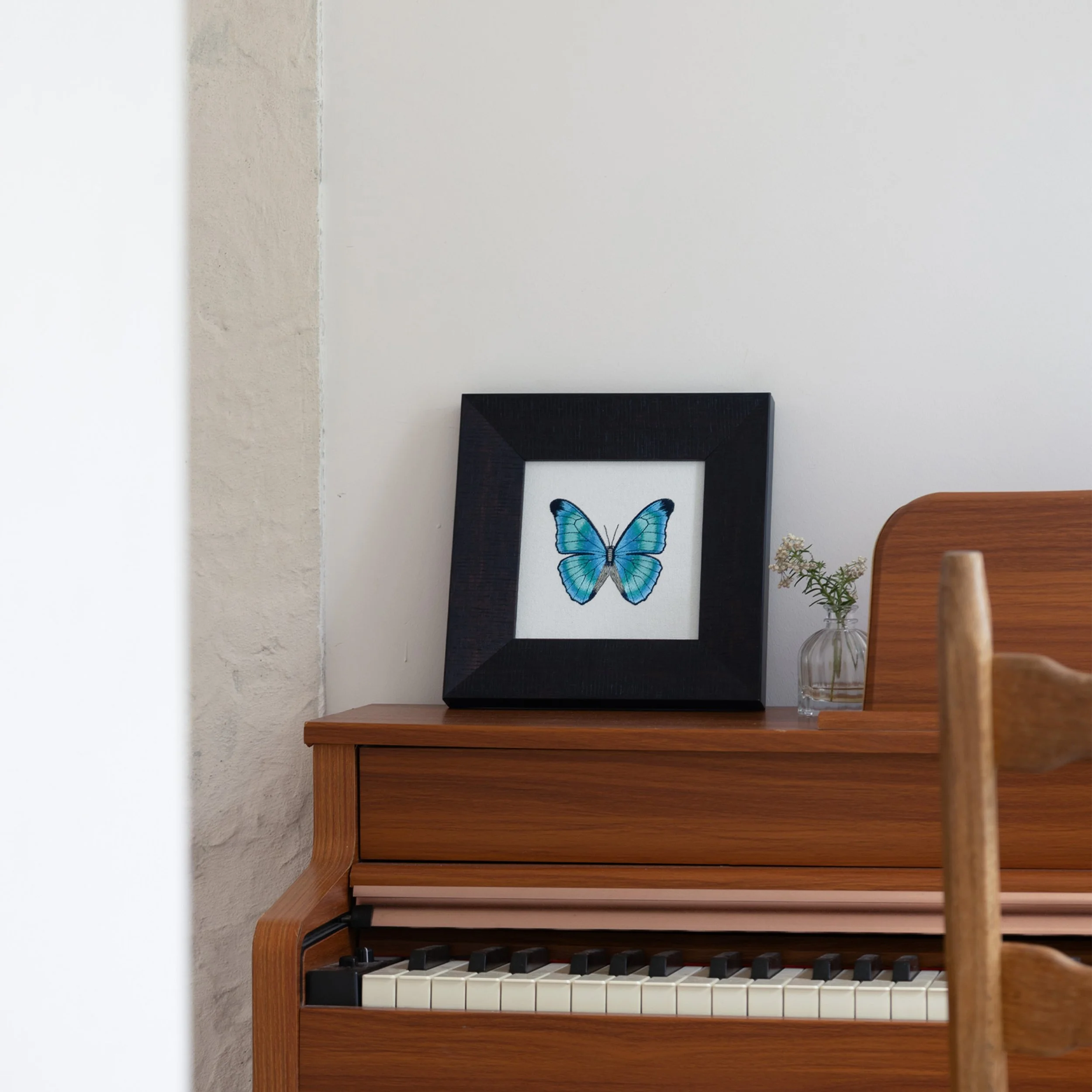 Aetheria butterfly embroidered wall art displayed on a wooden piano with a small glass vase.