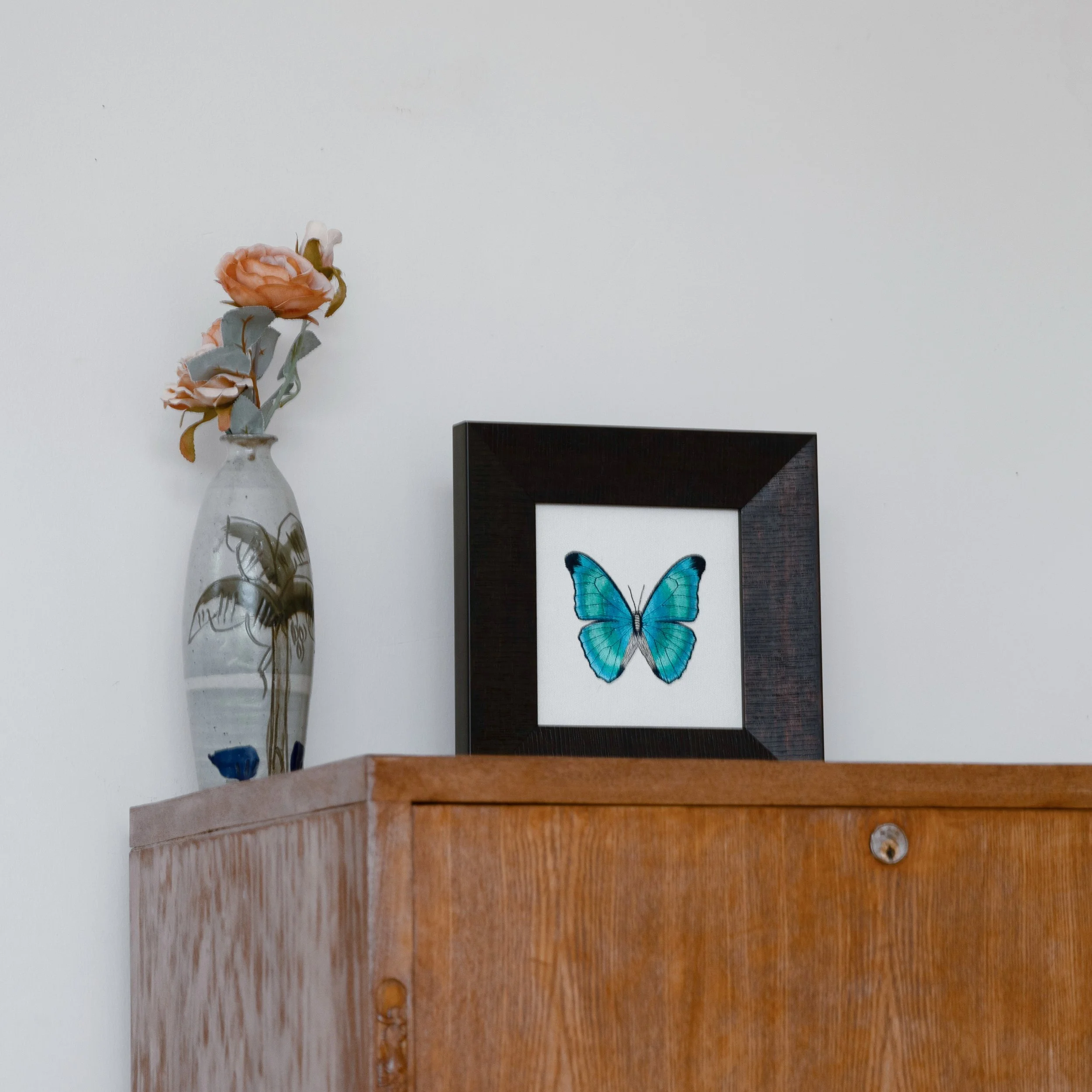 Aetheria butterfly embroidered wall art displayed on a wooden cabinet beside a ceramic vase with peach roses.