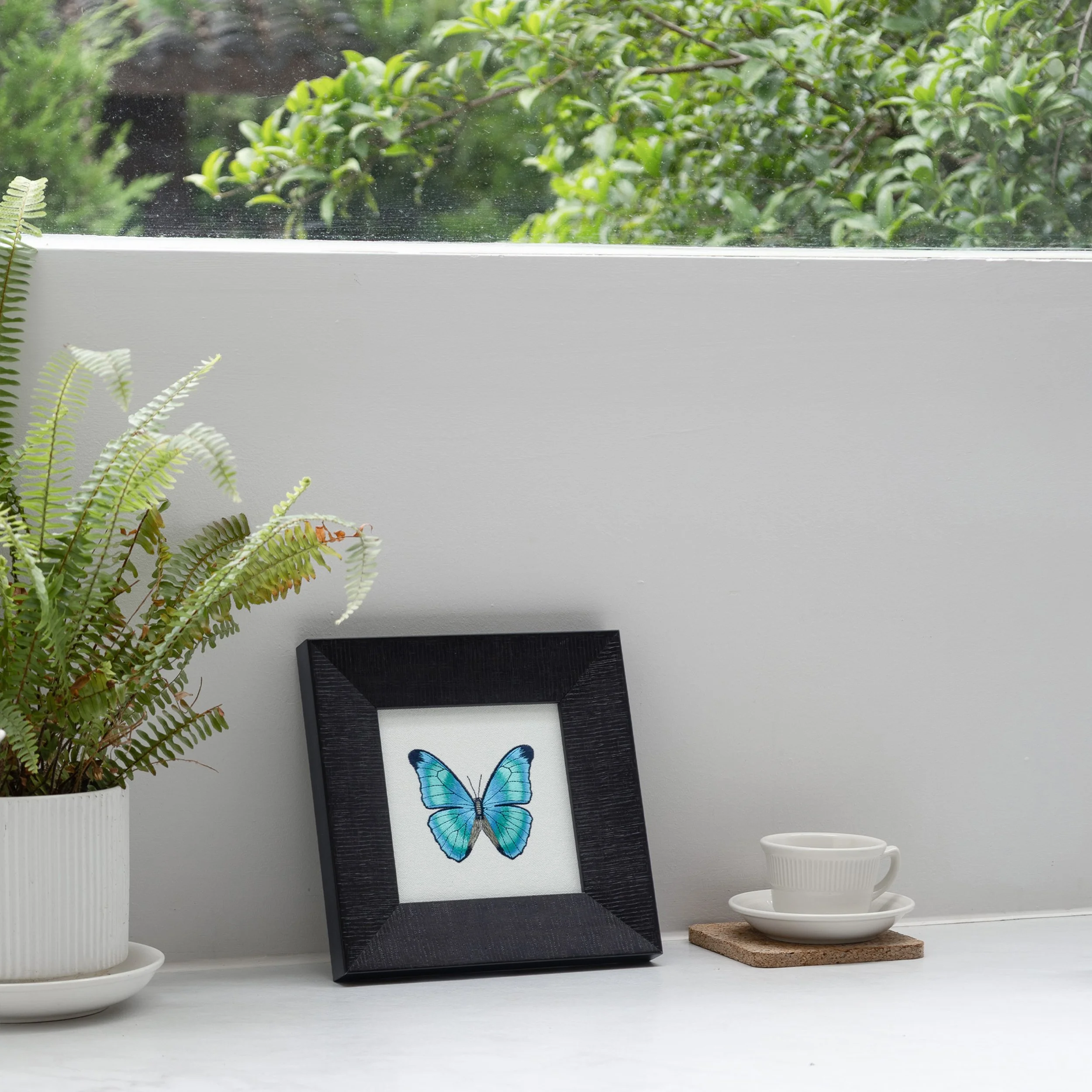 Framed teal butterfly embroidery leaning on a white countertop beside a fern and a white coffee cup.