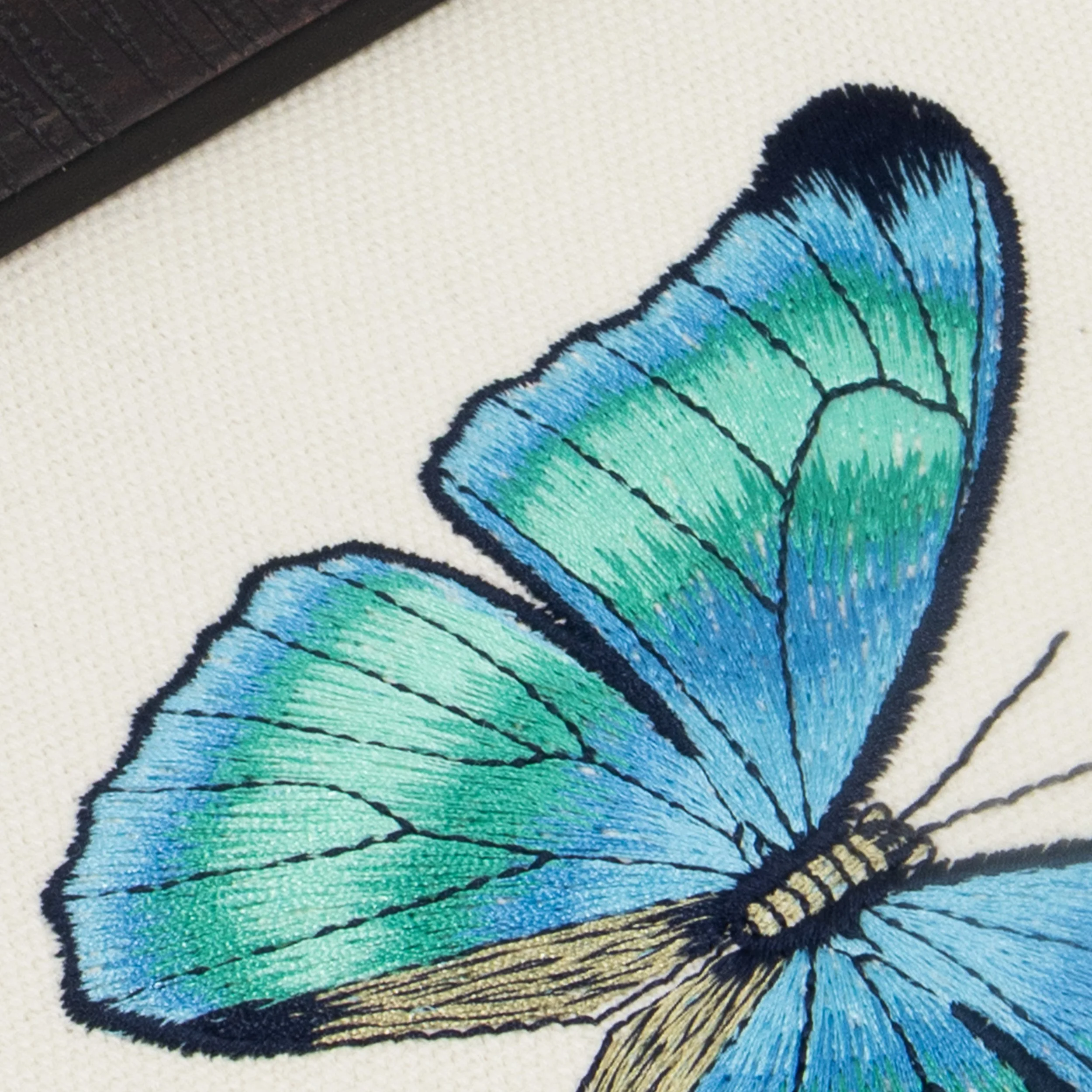 Close-up of Aetheria butterfly embroidery showing refined fine-line stitching and gradual teal-to-blue color transitions.