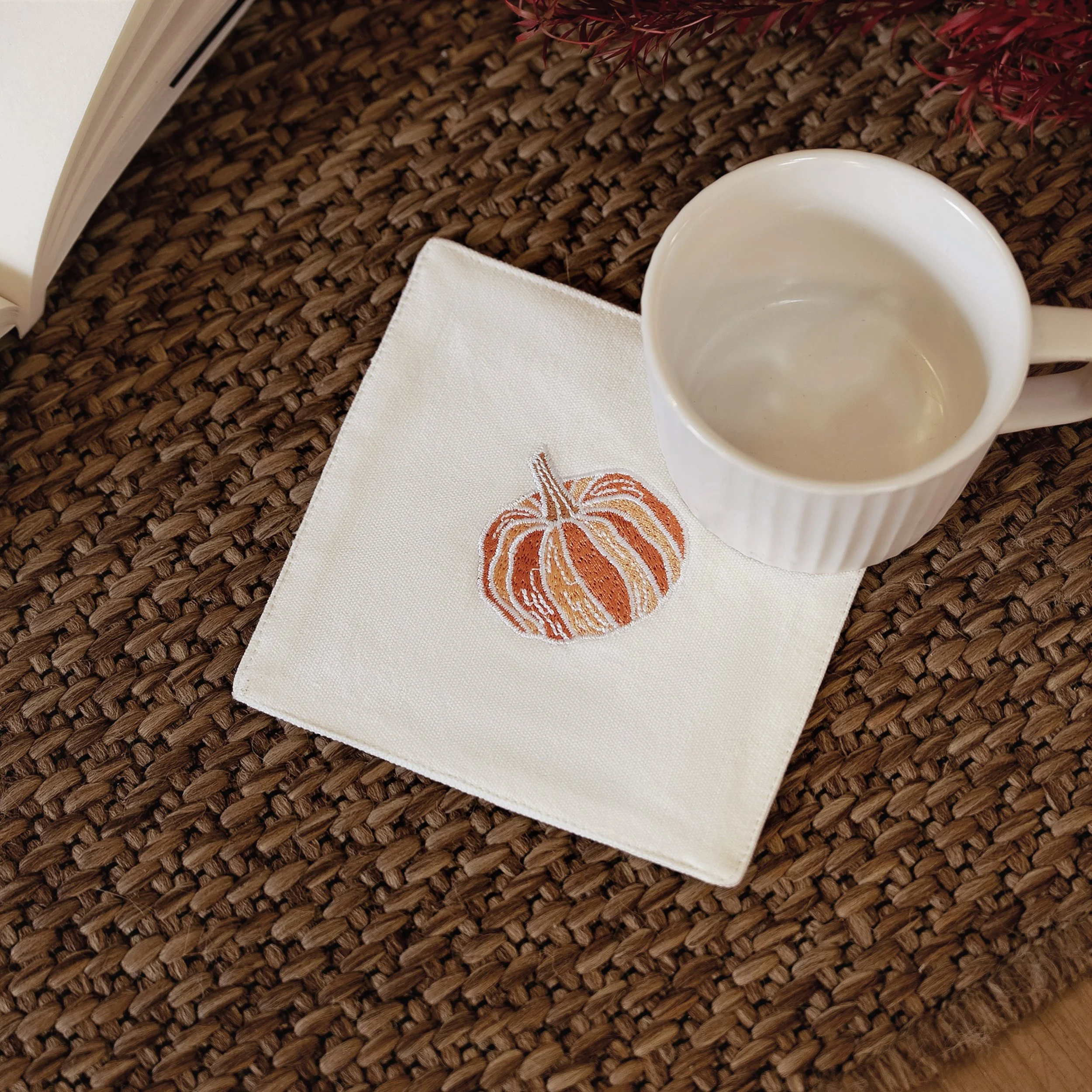 Pumpkin-embroidered coaster placed on a woven brown mat with a white mug — cozy fall table setup by Pam Pan, autumn home decoration and coffee coaster idea.