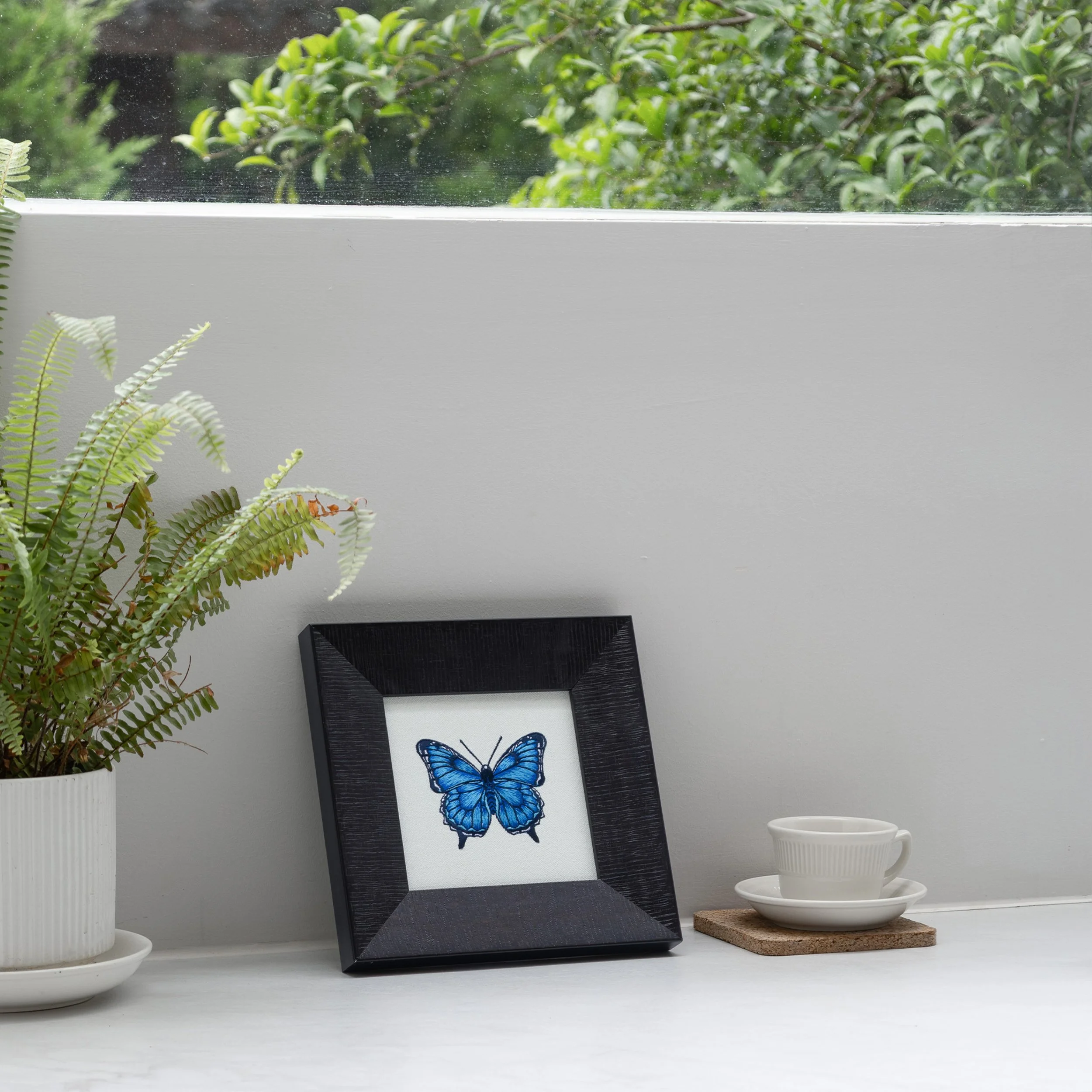 Blue butterfly embroidered wall art in a dark wood frame displayed on a white countertop beside a coffee cup and potted fern