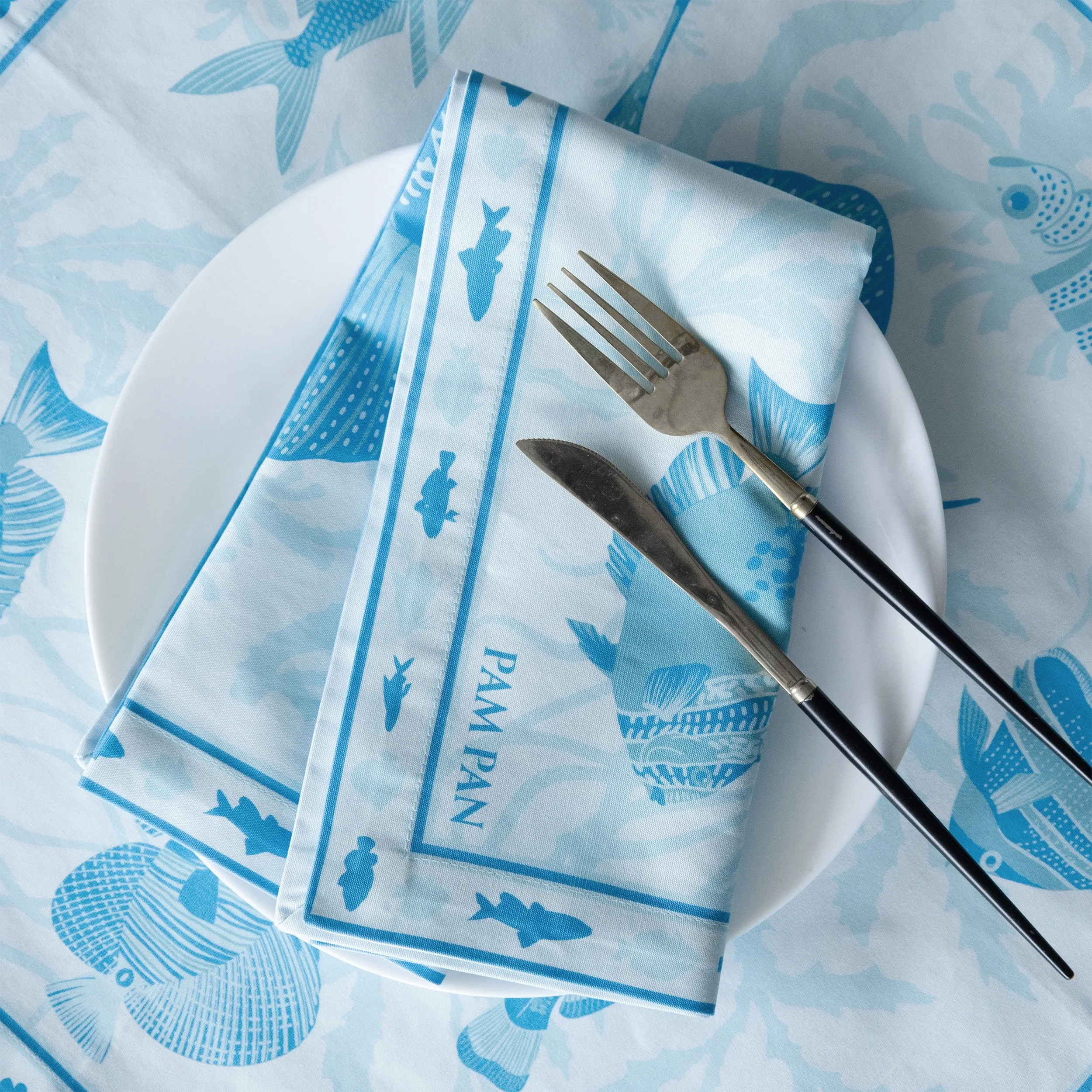 Aqua printed napkin paired with white plate and silver cutlery. Double-sided cotton napkin with ocean-inspired fish pattern by Pam Pan.