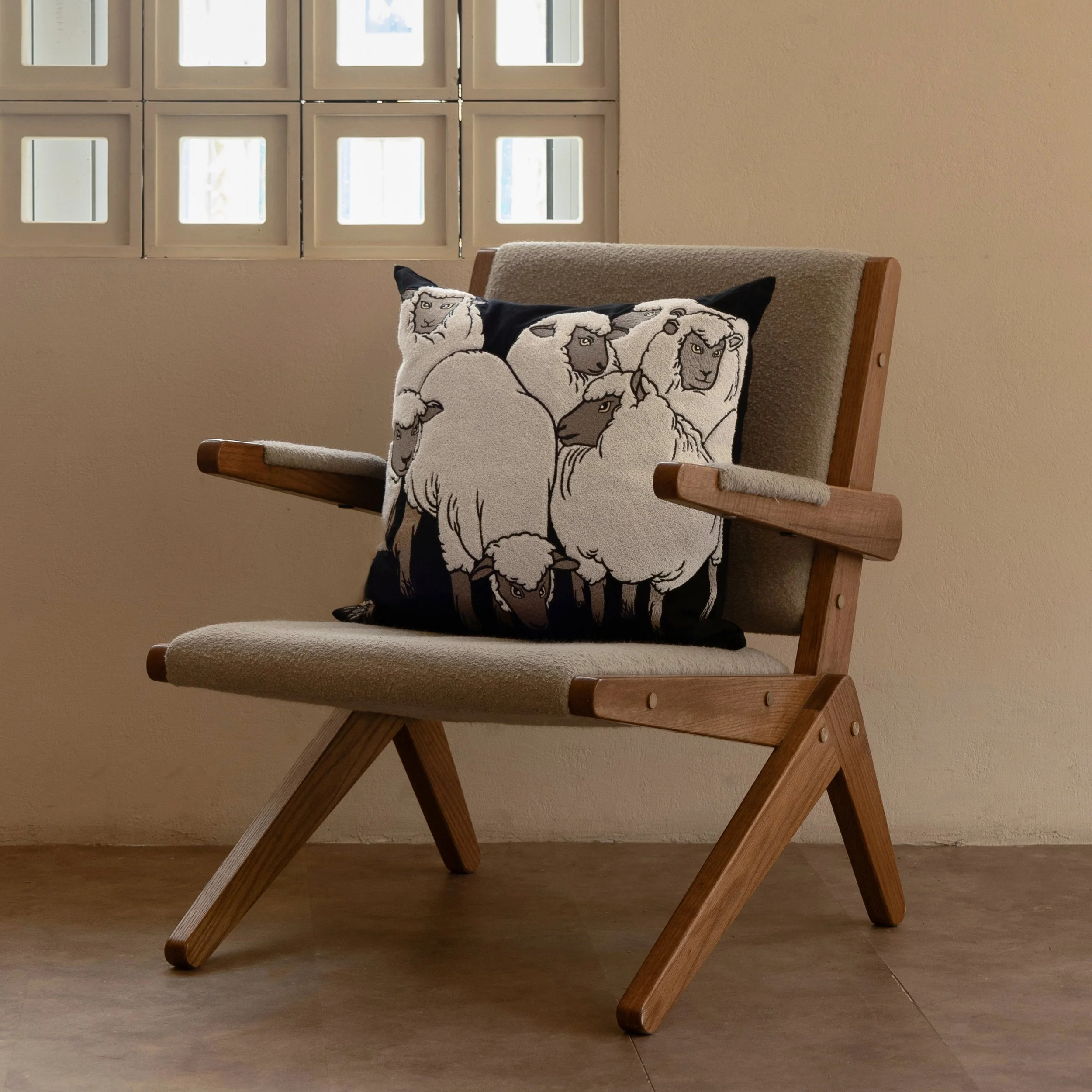 The Flock sheep embroidered throw pillow displayed on a modern wooden lounge chair