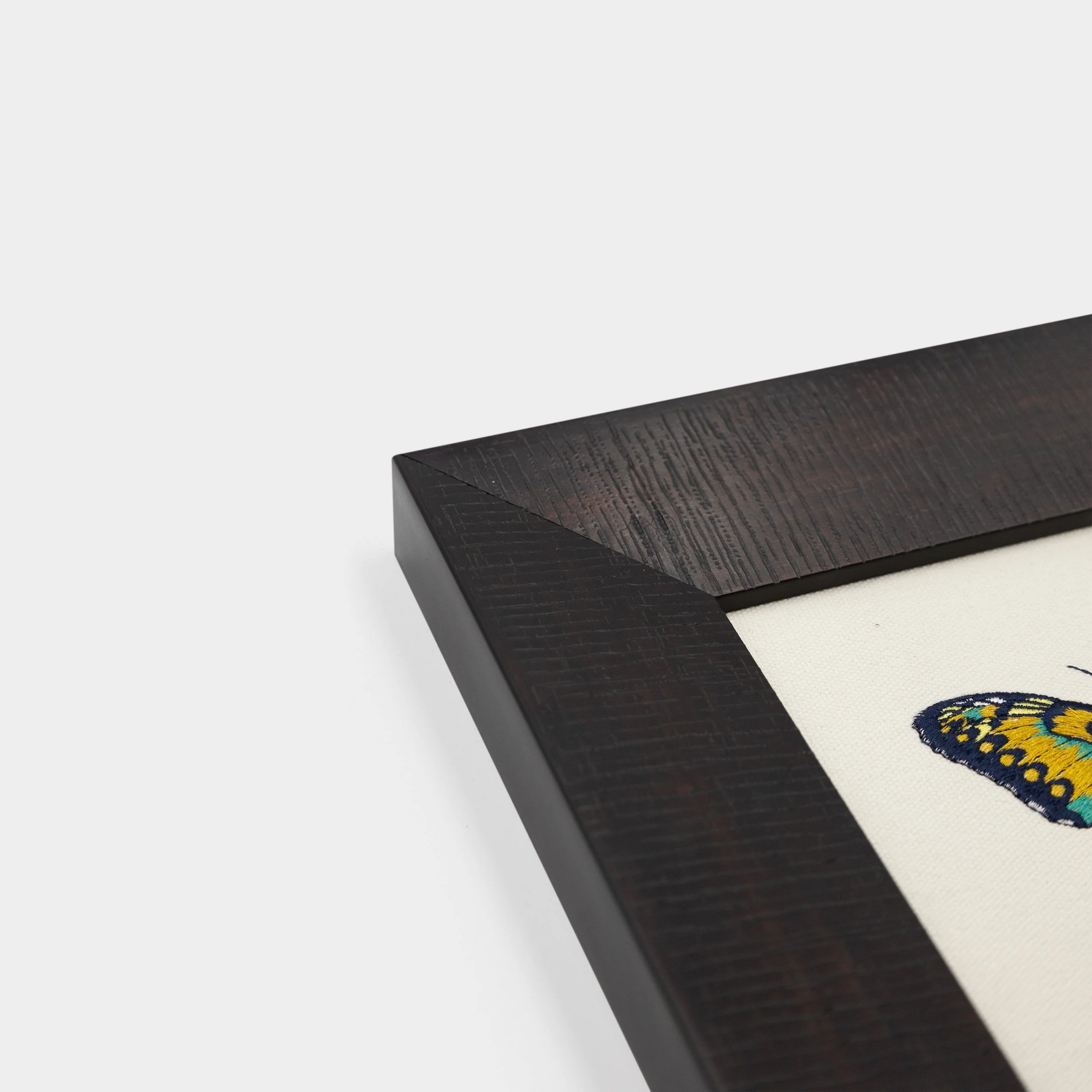 Corner detail of the dark solid wood frame surrounding the Ignisia butterfly embroidered artwork.
