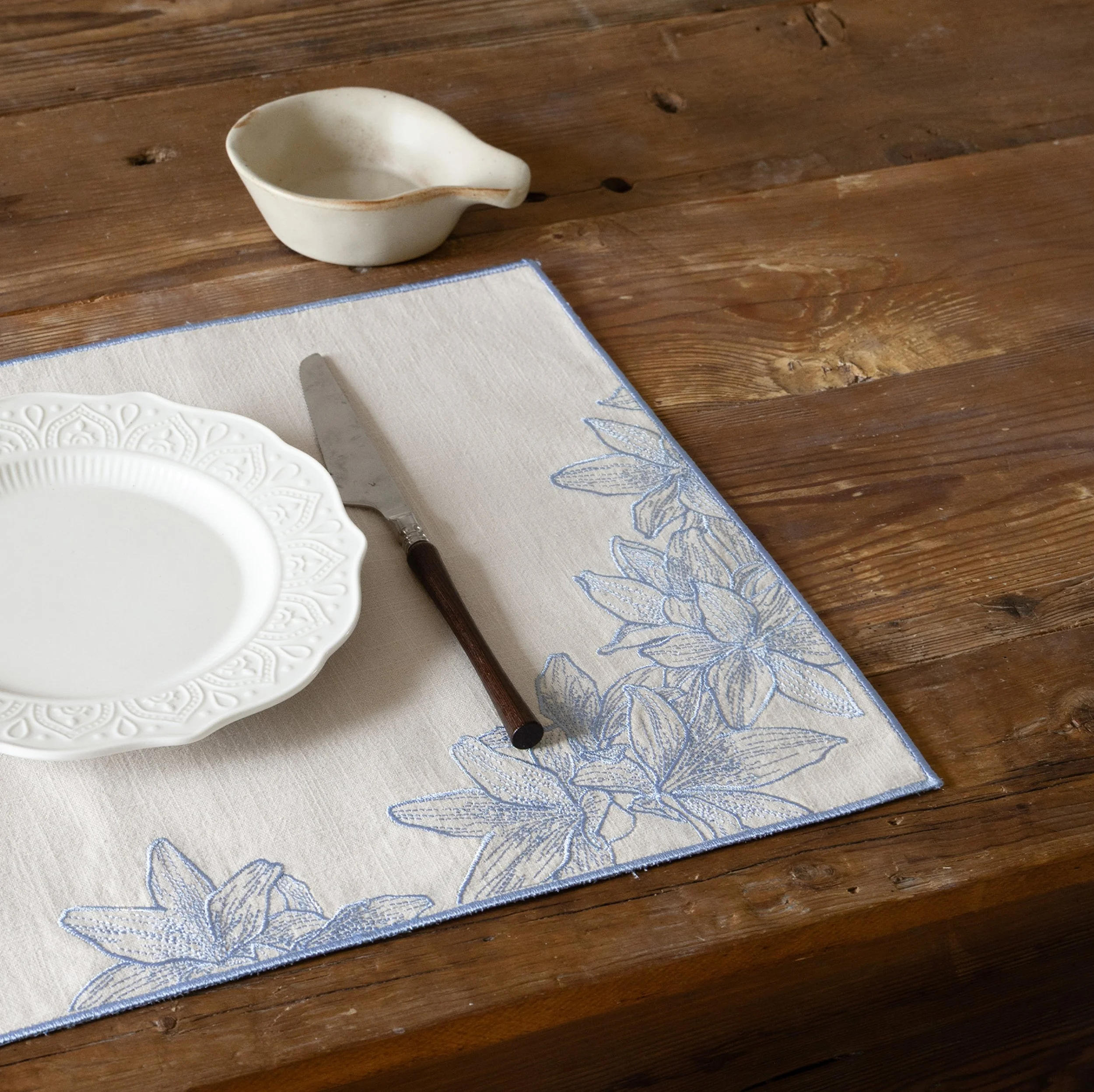 Blue lily embroidered placemat styled on a rustic wooden dining table with a white ceramic plate and wooden-handle knife