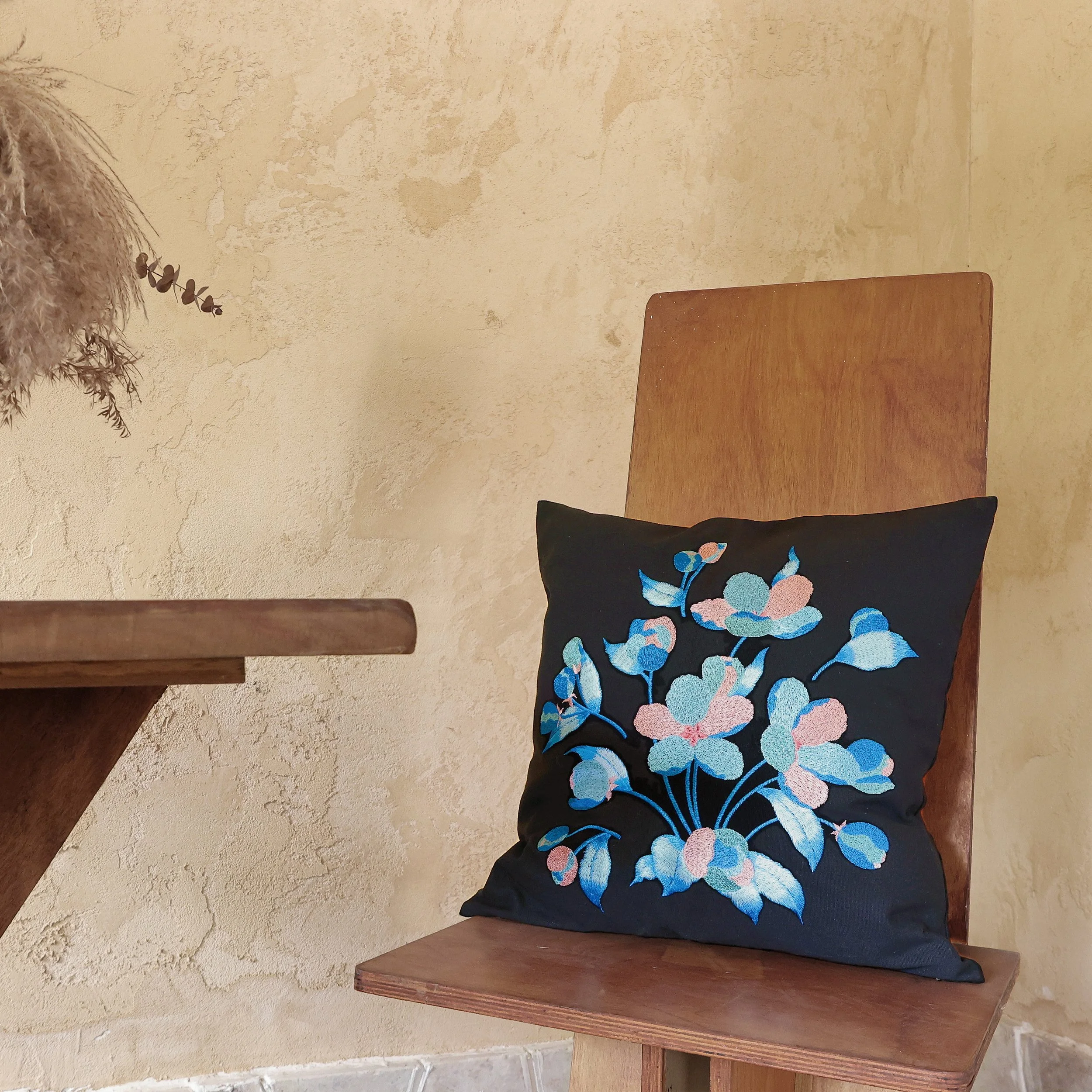 Black embroidered floral cushion from the Etherelle Series placed on a wooden chair in a neutral home setting.