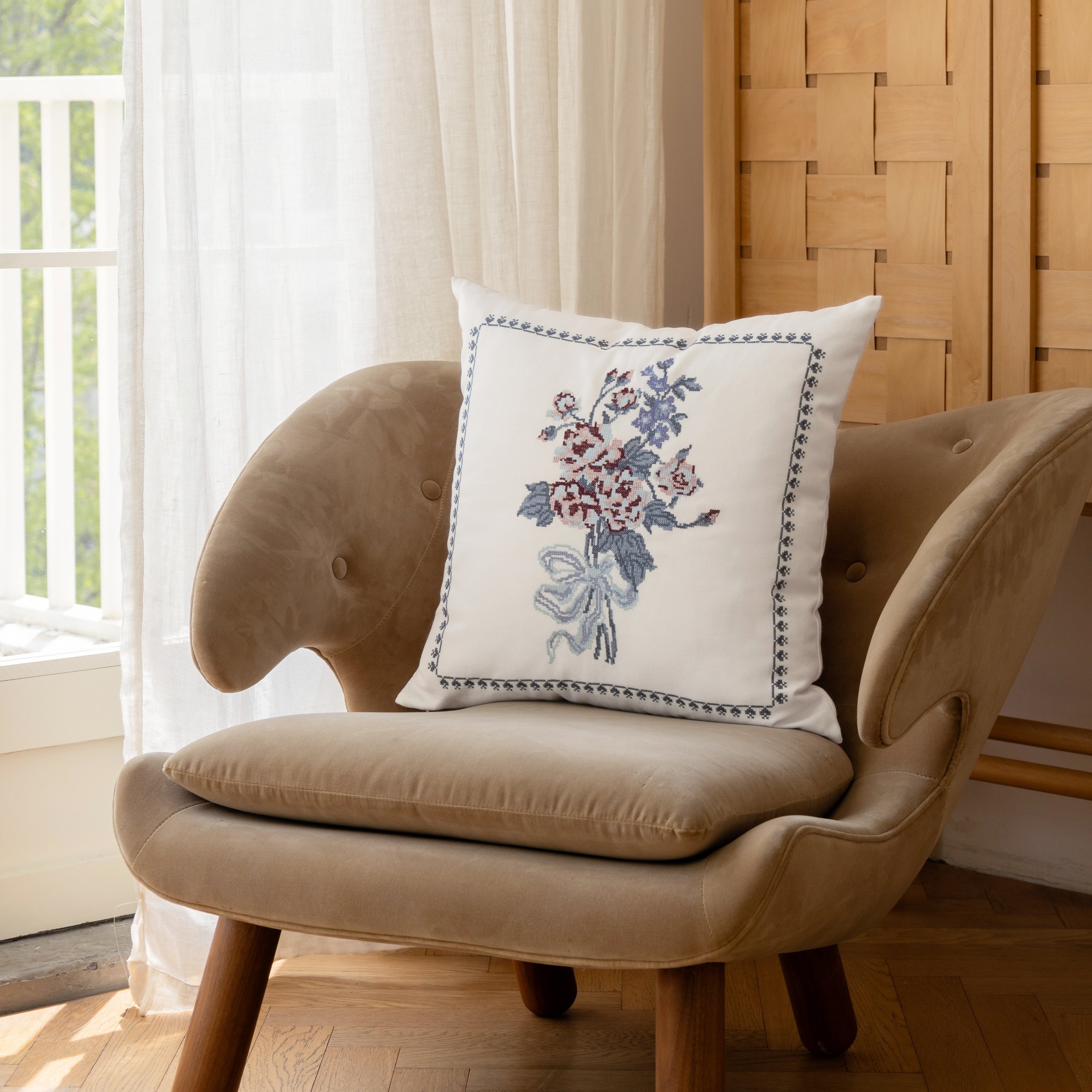 Bouquet cross stitch embroidered throw pillow styled on a suede armchair beside sheer curtains and natural light
