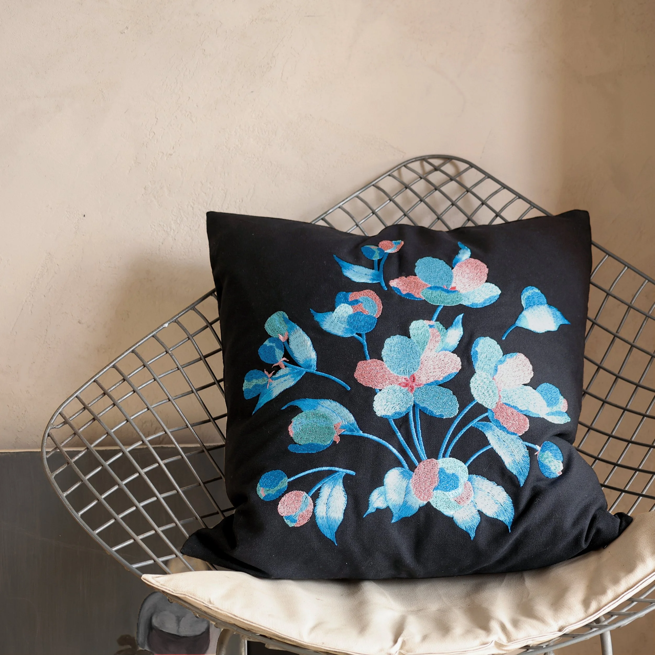 Etherelle floral embroidered pillow in black displayed on a modern wire chair, blending craft and contemporary style.