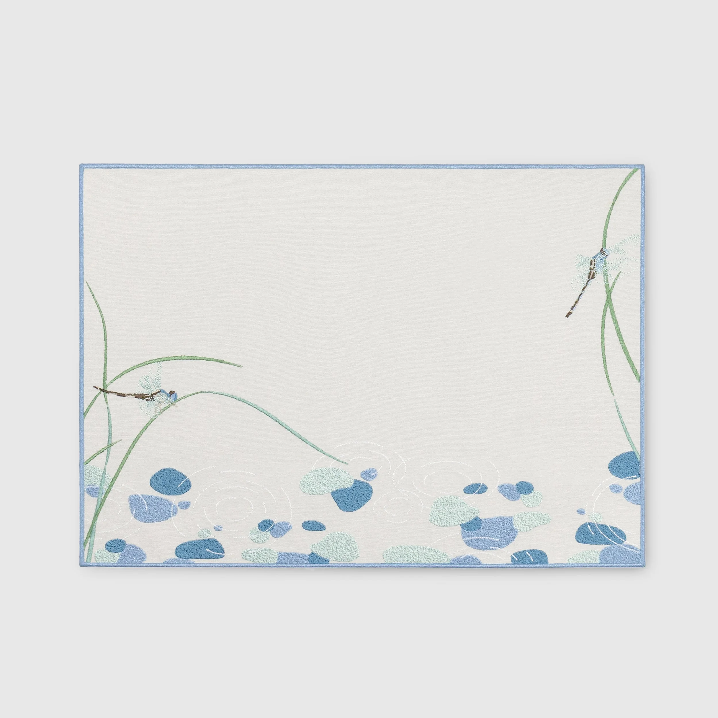 Riparia embroidered placemat featuring dragonflies, reeds, water ripples, and blue stone motifs on a neutral background