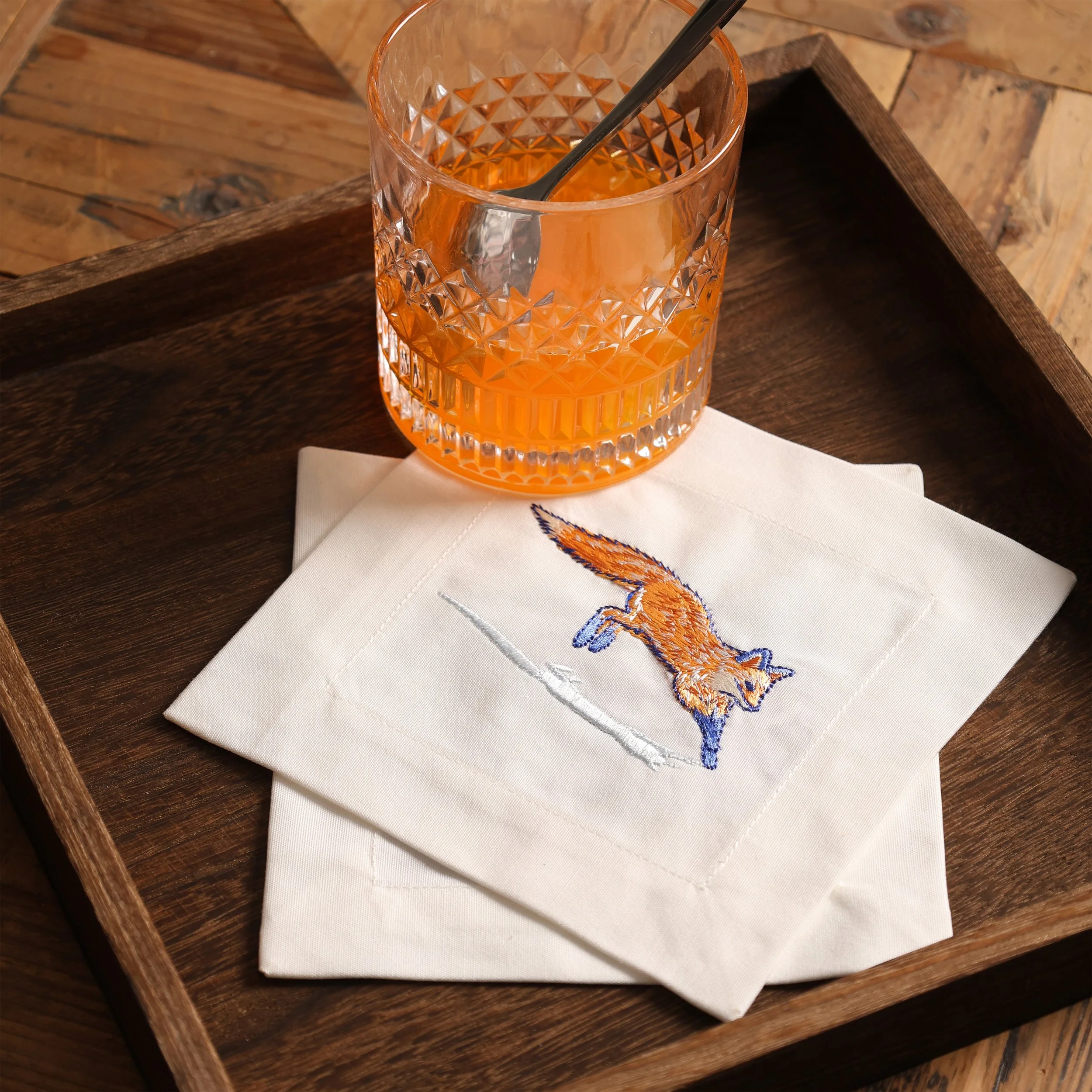 Fox embroidered cocktail napkin placed in a wooden tray with an orange drink glass
