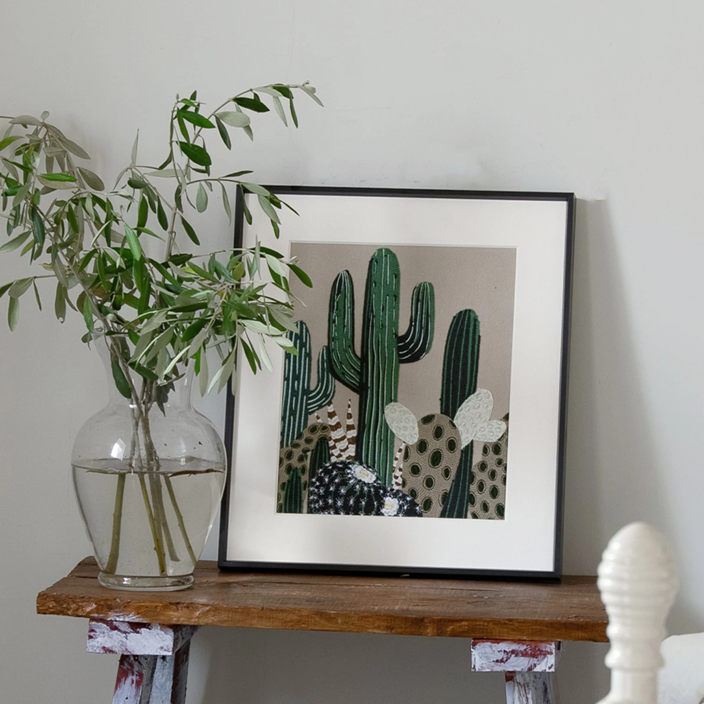 Framed cactus embroidery wall art displayed on a wooden bench beside a glass vase with olive branches