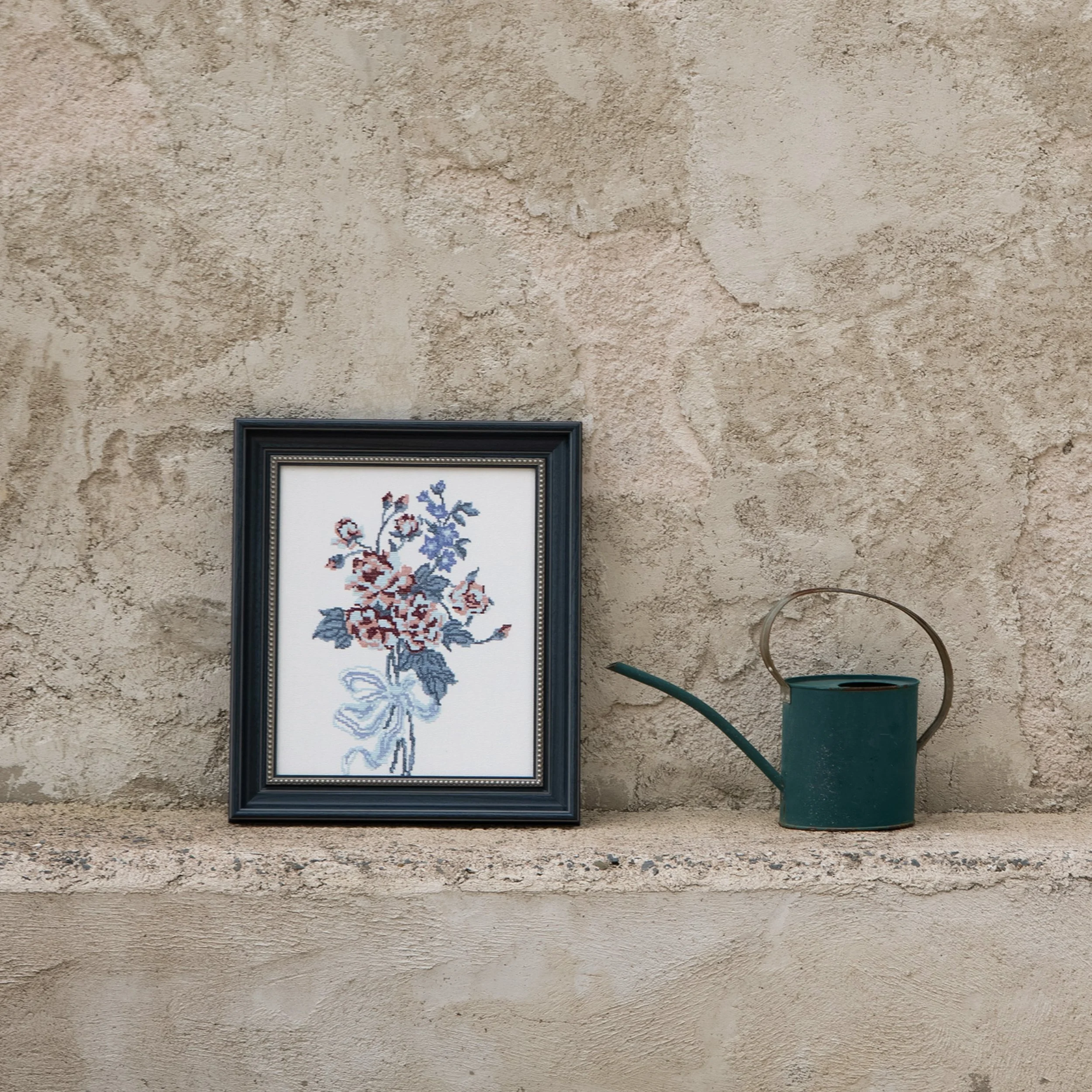 Cross stitch floral wall art in a navy frame displayed against a textured stone wall