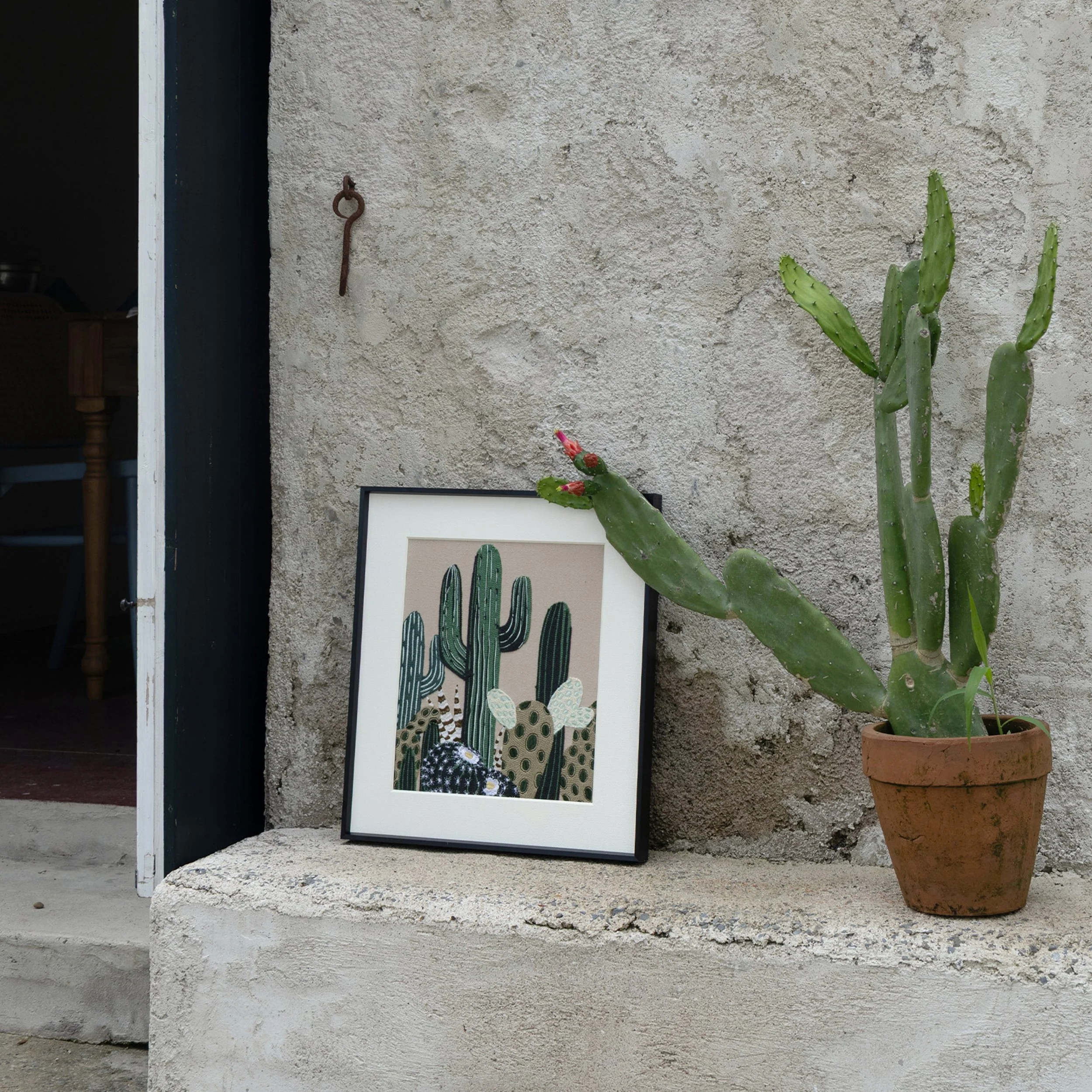Framed cactus embroidery artwork placed outdoors beside a potted cactus