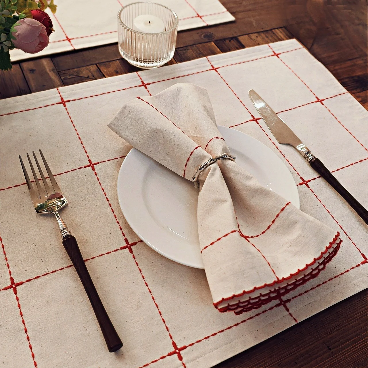 Red grid embroidered light beige cotton placemat in a table setting with the matching napkin on top of a plate.