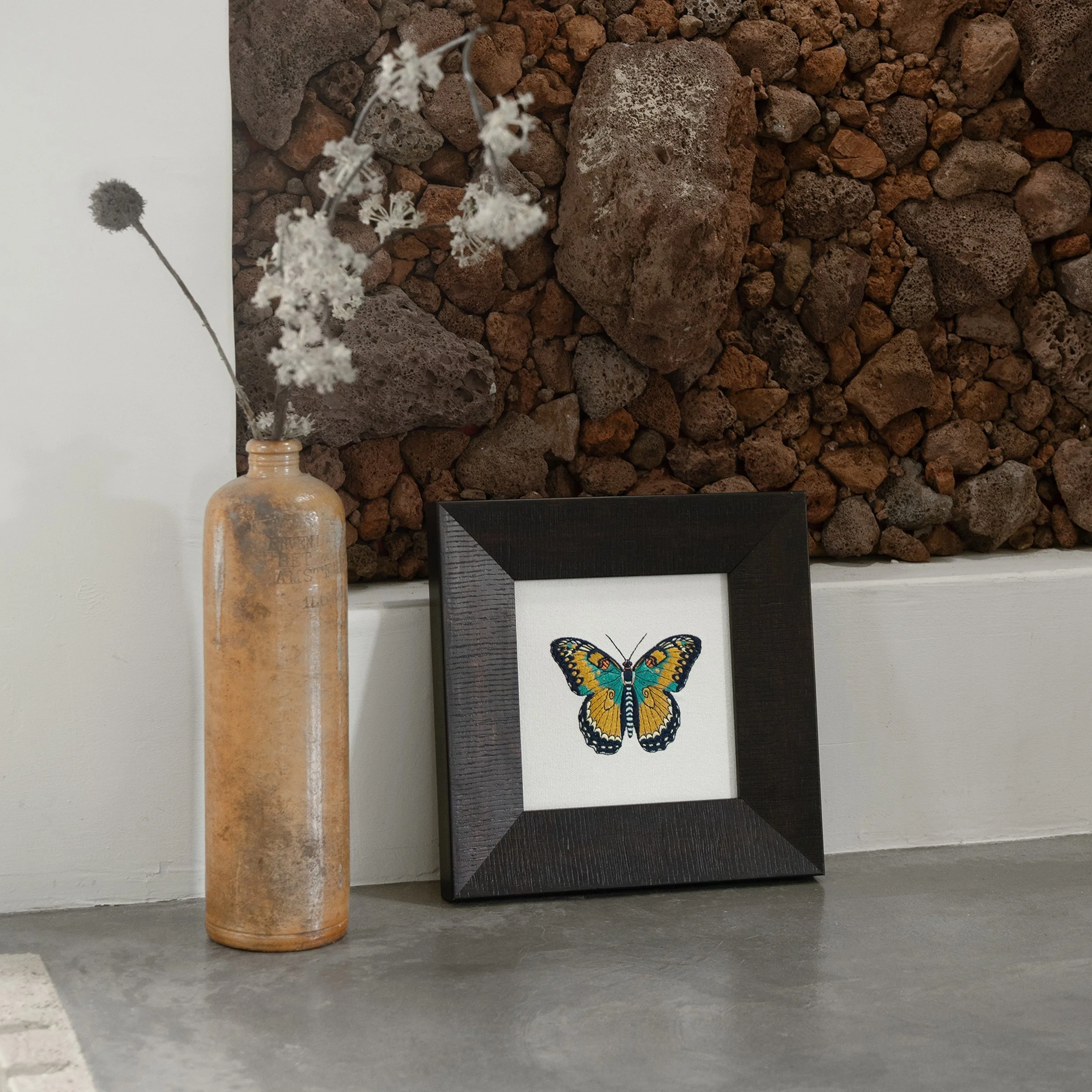 Ignisia butterfly embroidered wall art displayed beside a ceramic bottle against a textured stone wall backdrop.