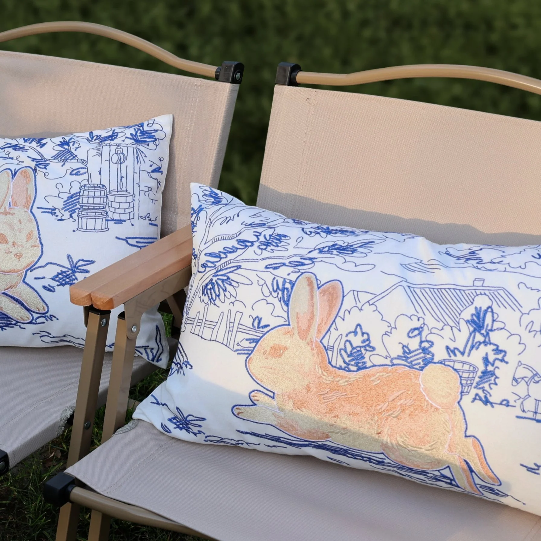 Whimsy Series embroidered lumbar pillow with a leaping rabbit and blue pastoral landscape pattern, set on beige outdoor chairs under natural light.