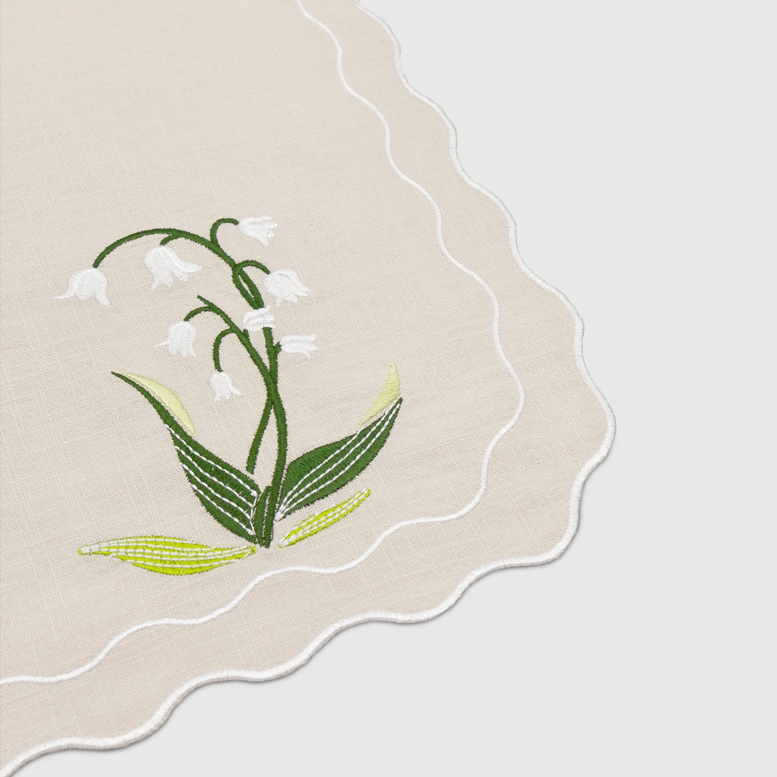 Close-up detail of lily of the valley embroidery on a scalloped-edge napkin showing white flowers and green stitched leaves