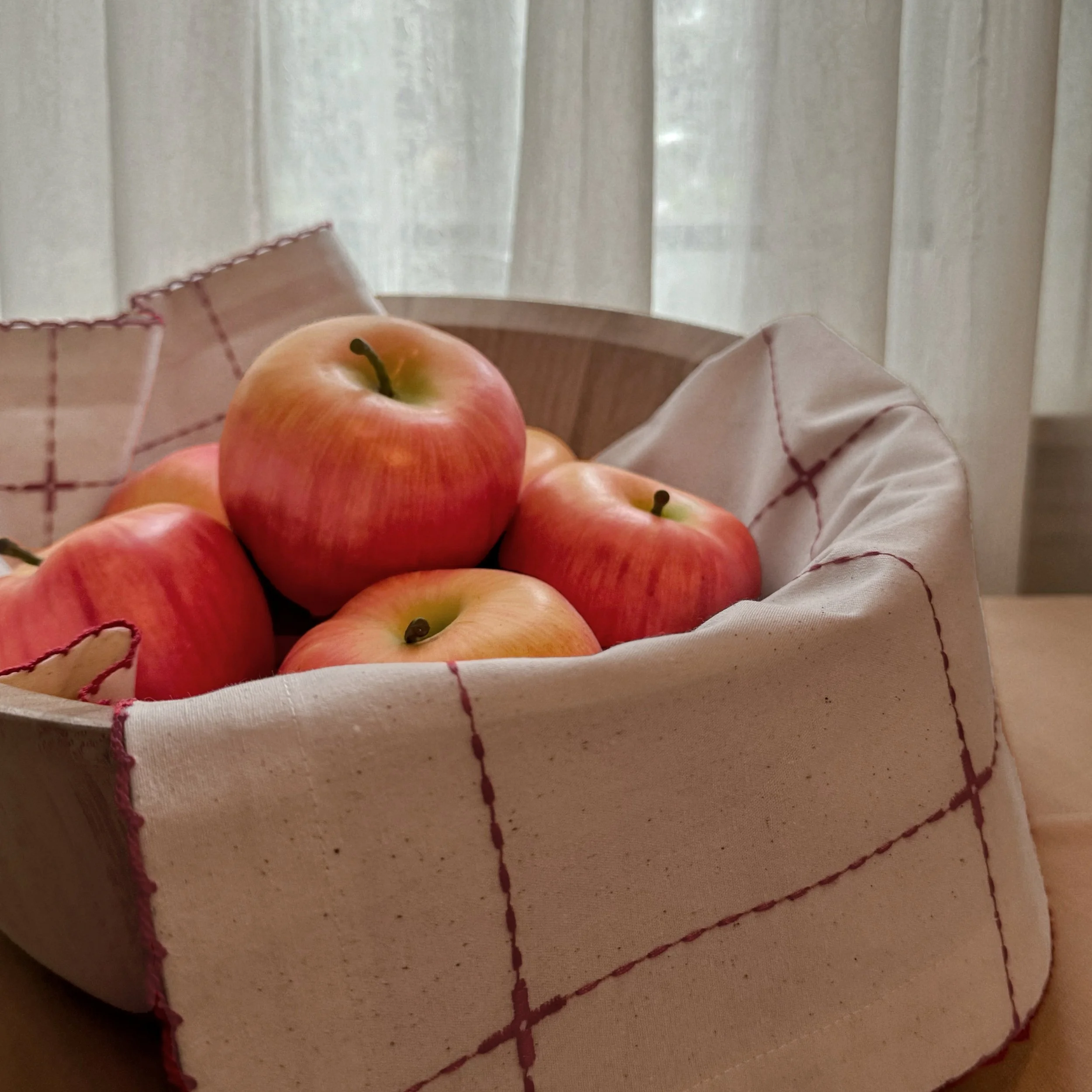 Scallop-edged cotton napkin with embroidered grid details in a wooden basket with apples on top.