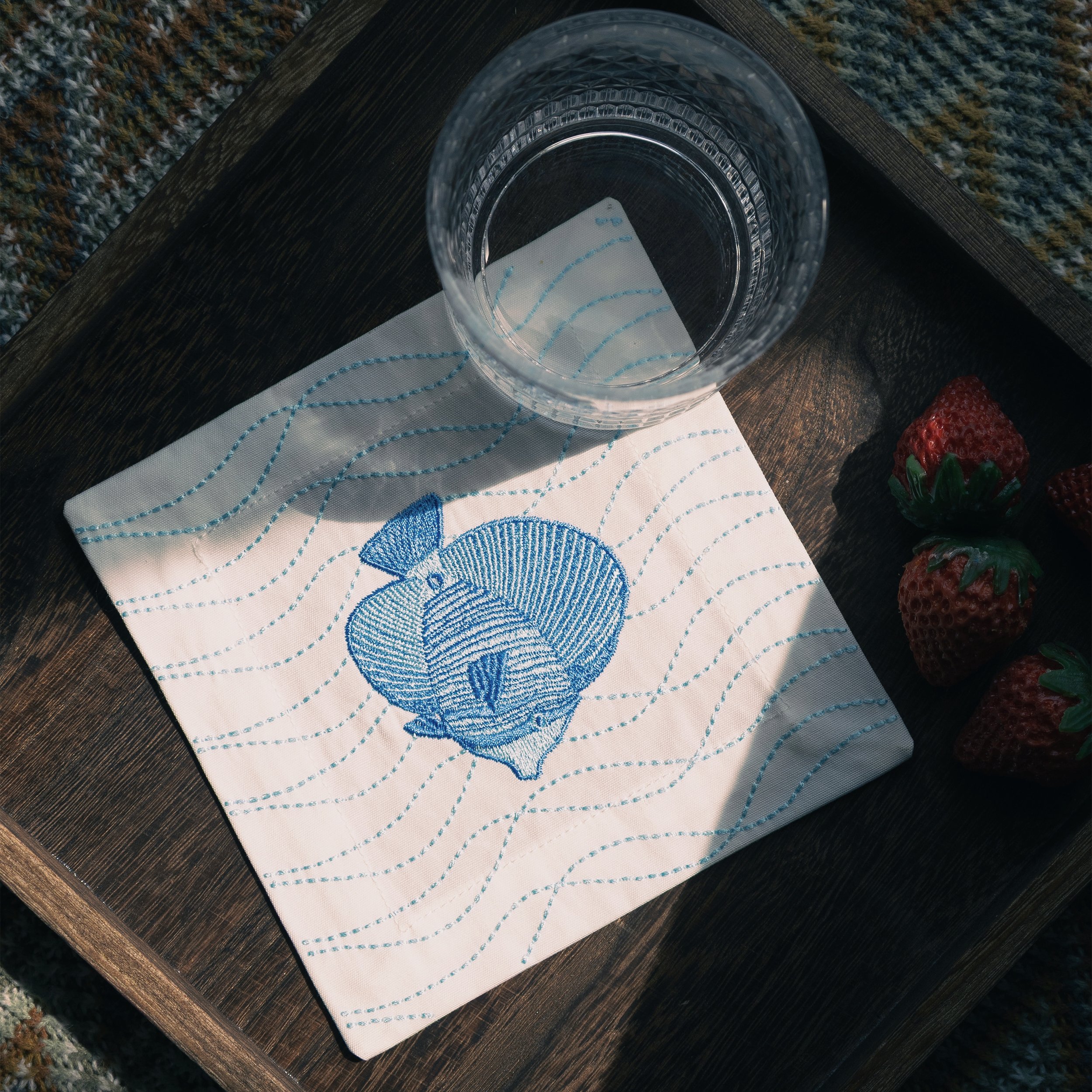 Blue fish embroidered cocktail napkin placed in a wooden serving tray with a glass and fruits — ocean-inspired cotton coaster by Pam Pan.
