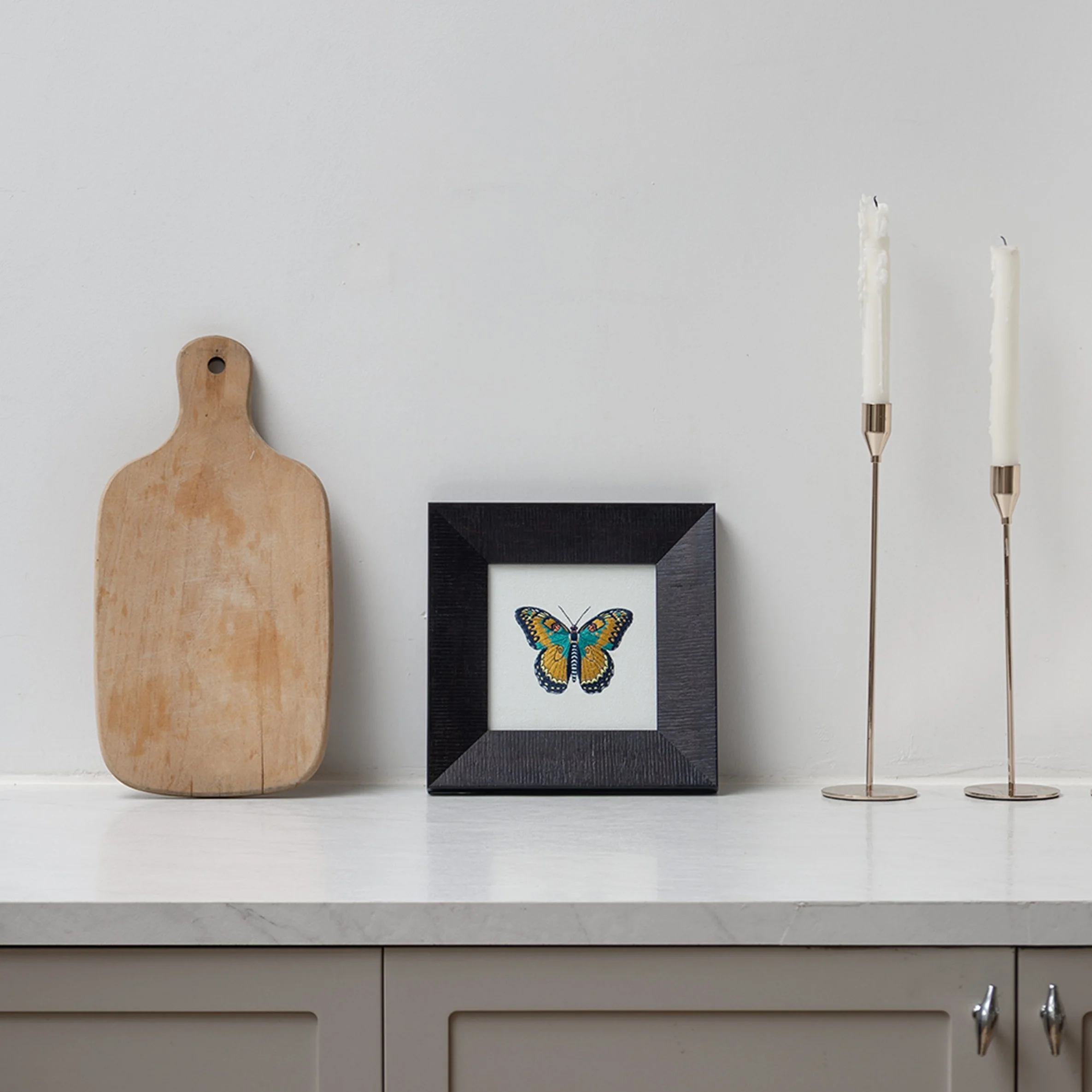 Ignisia butterfly embroidered wall art styled on a white countertop beside a wooden cutting board and candle holders.