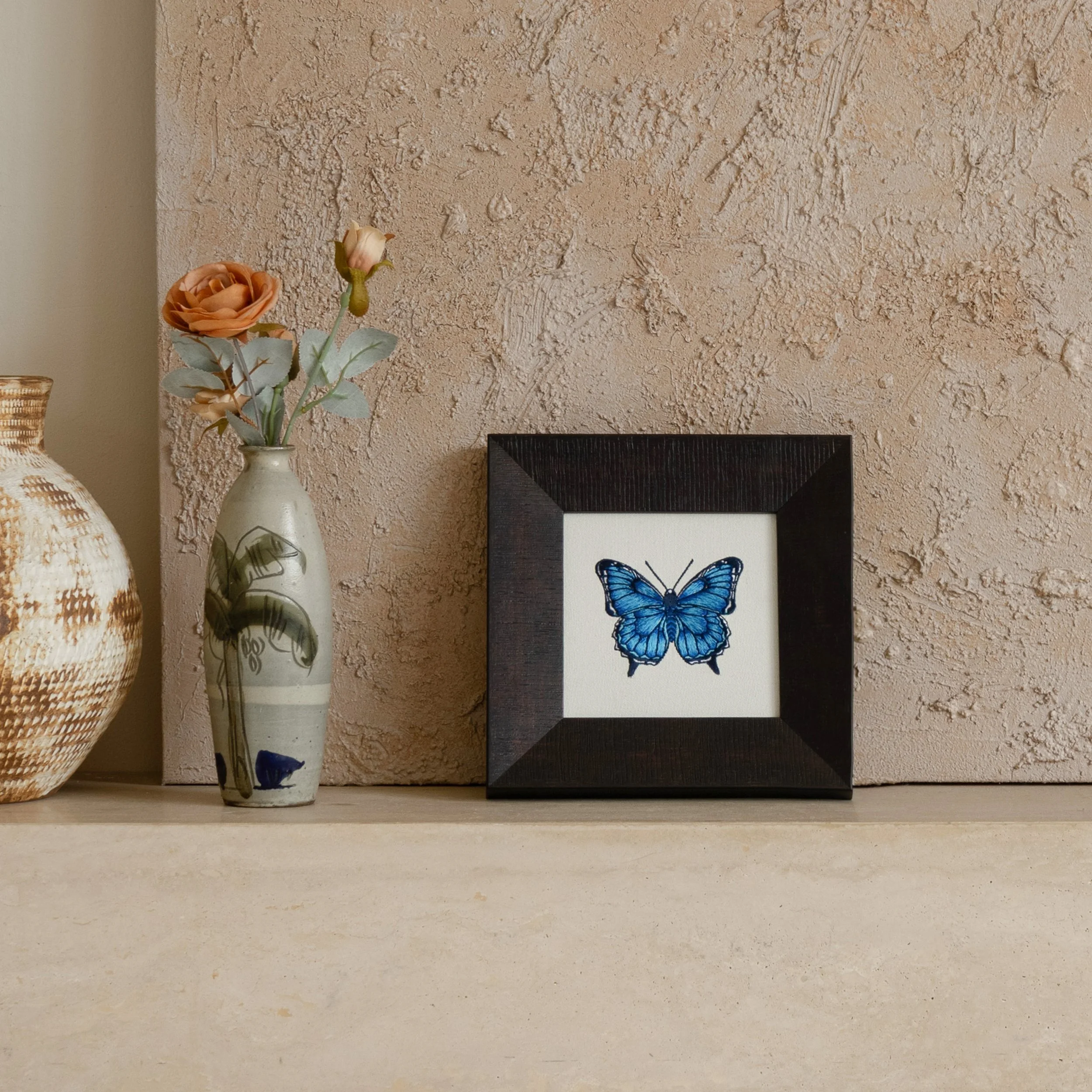Framed blue butterfly embroidery displayed on a shelf beside a ceramic vase against a textured wall