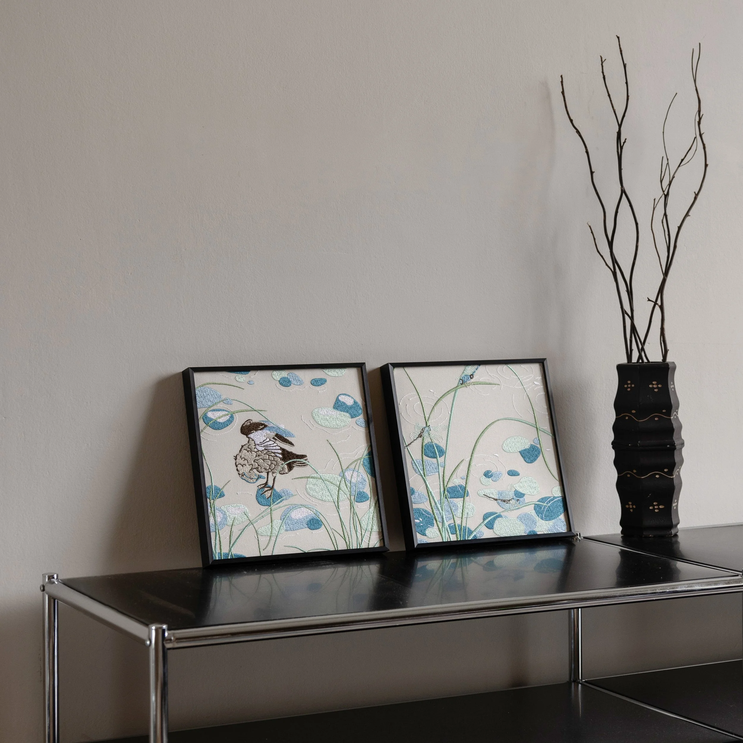 Two framed embroidered artworks from the Riparia collection displayed on a modern black metal console table, featuring a duck and dragonfly pond scene.