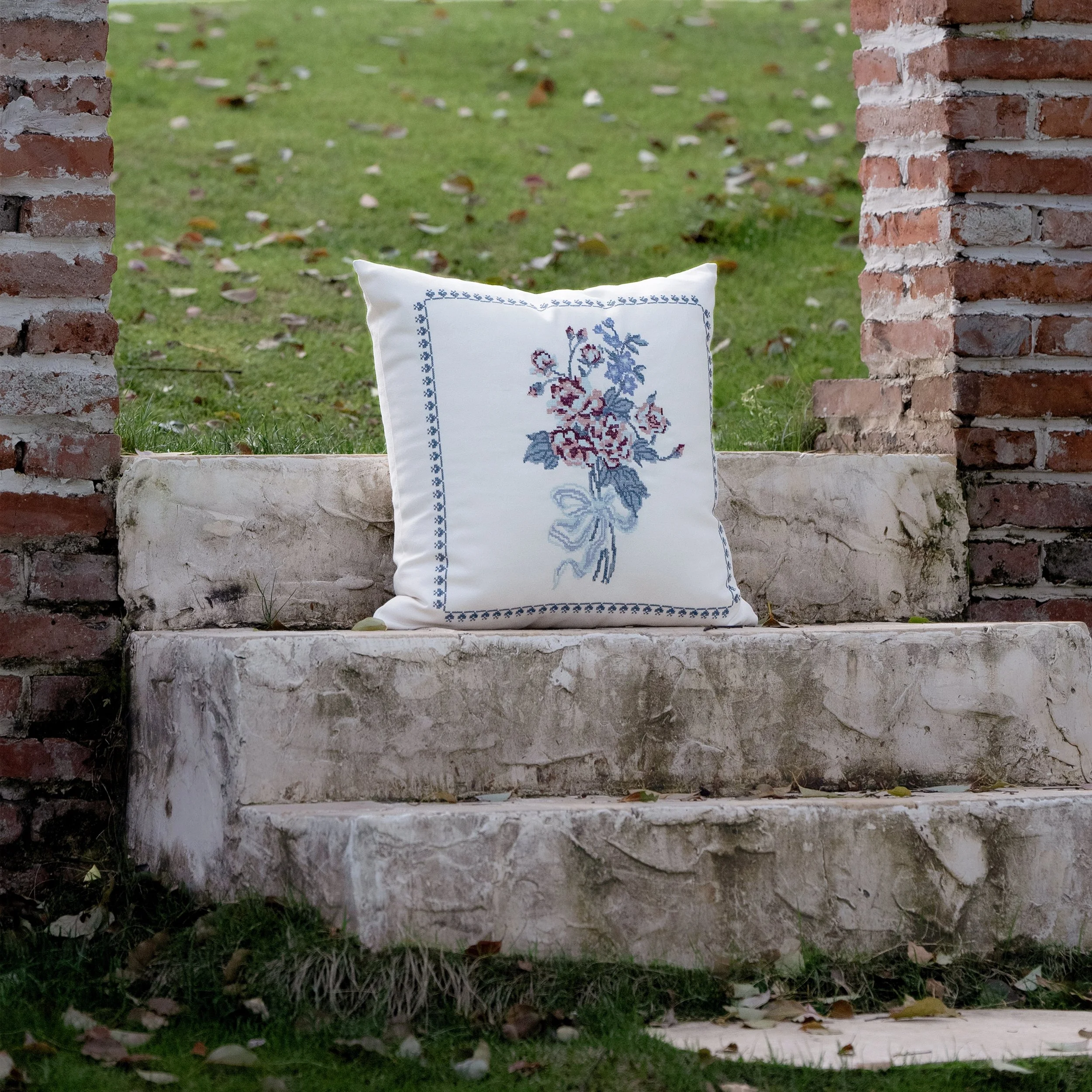 Bouquet floral cross stitch embroidered throw pillow placed on weathered stone steps surrounded by a garden setting