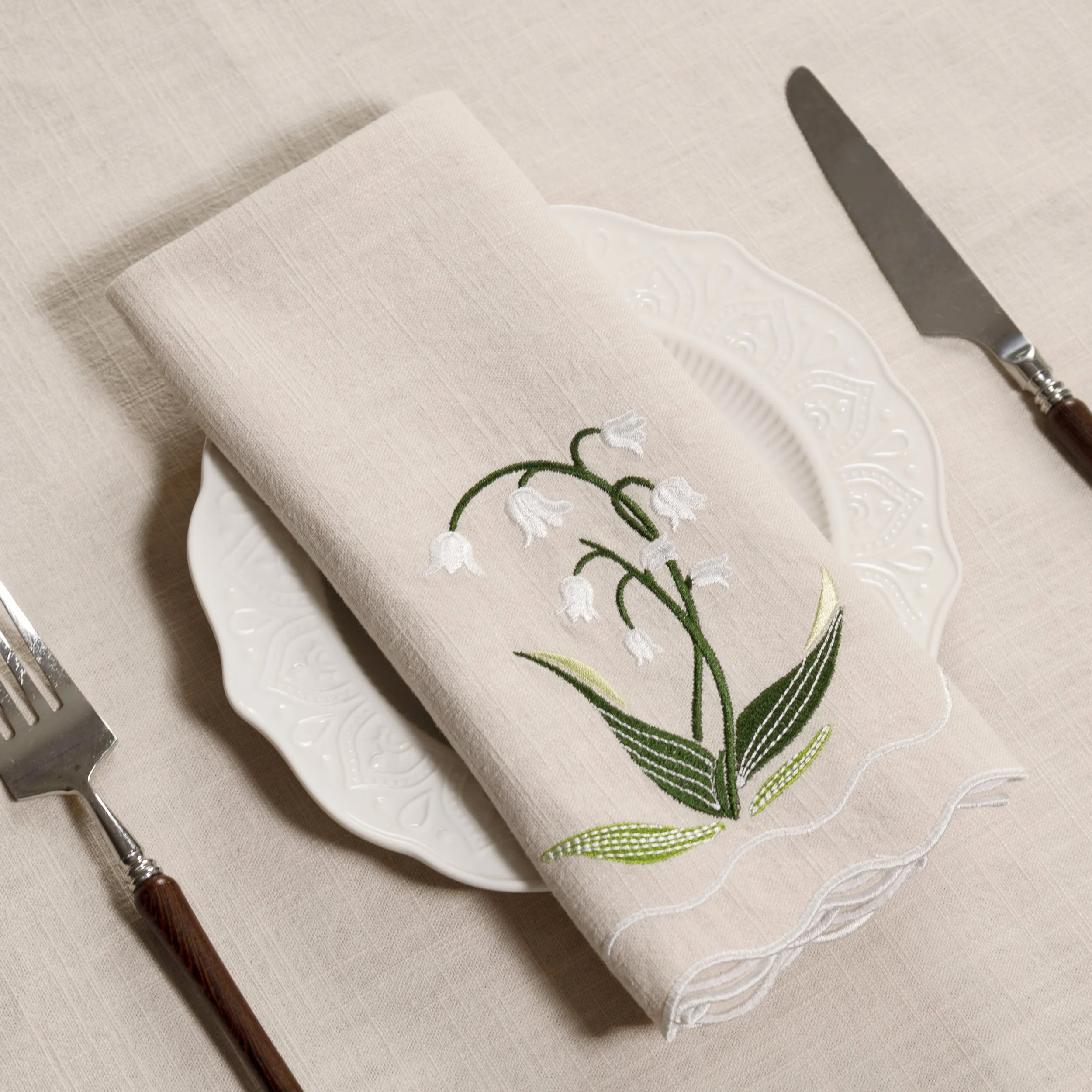 Folded lily of the valley embroidered napkin displayed on a white dinner plate with botanical embroidery and scalloped edge detail