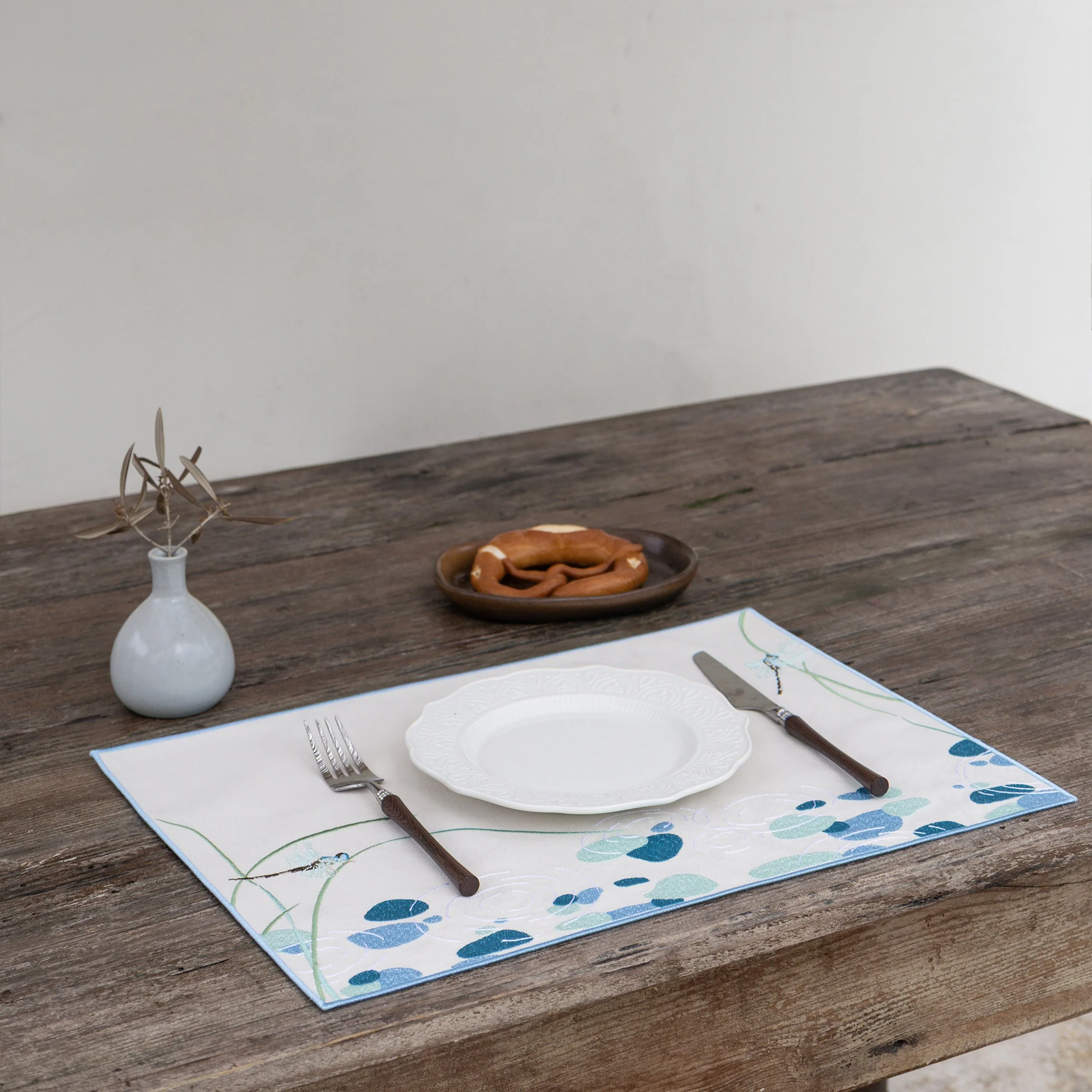 Riparia embroidered placemat styled on a rustic wooden dining table with a white ceramic plate and wooden-handle cutlery