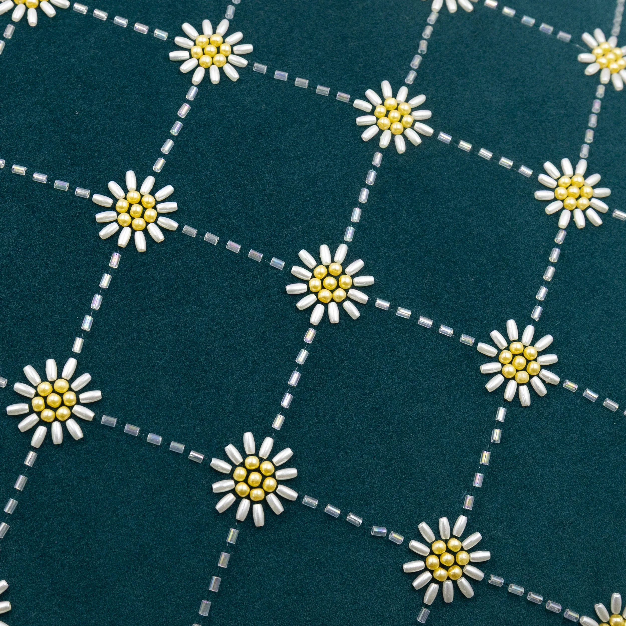 Close-up of daisy beading embroidery showing ivory petal beads, golden bead centers, and geometric lattice detailing on teal velvet fabric.