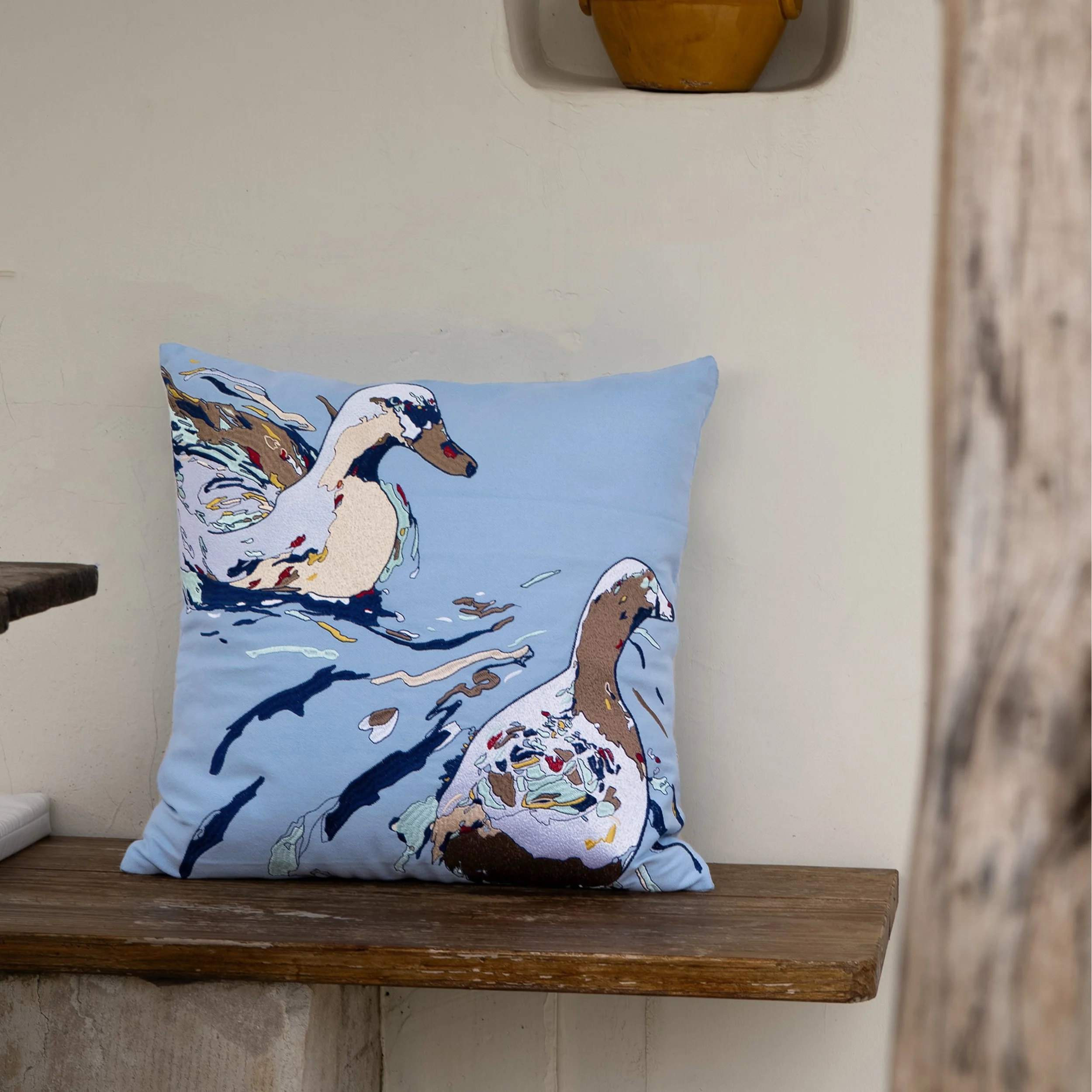 Drift embroidered throw pillow displayed on a rustic wooden bench, showcasing embroidered ducks and flowing water motifs in a calm, light-filled interior.