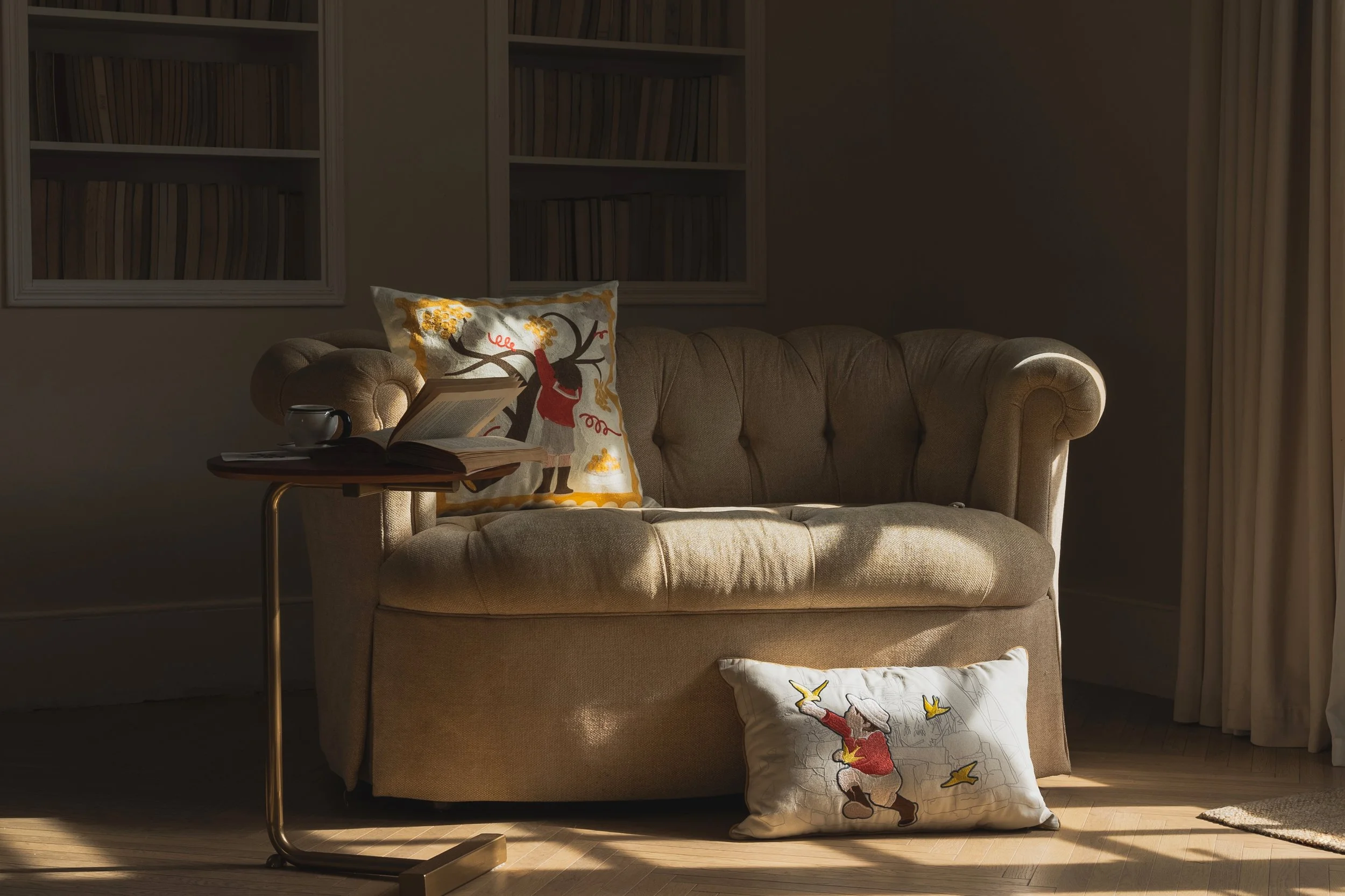 Embroidered throw pillow and lumbar pillow from the Wild Corner collection placed on a living room sofa, featuring child figure designs in a warm, sunlit interior.