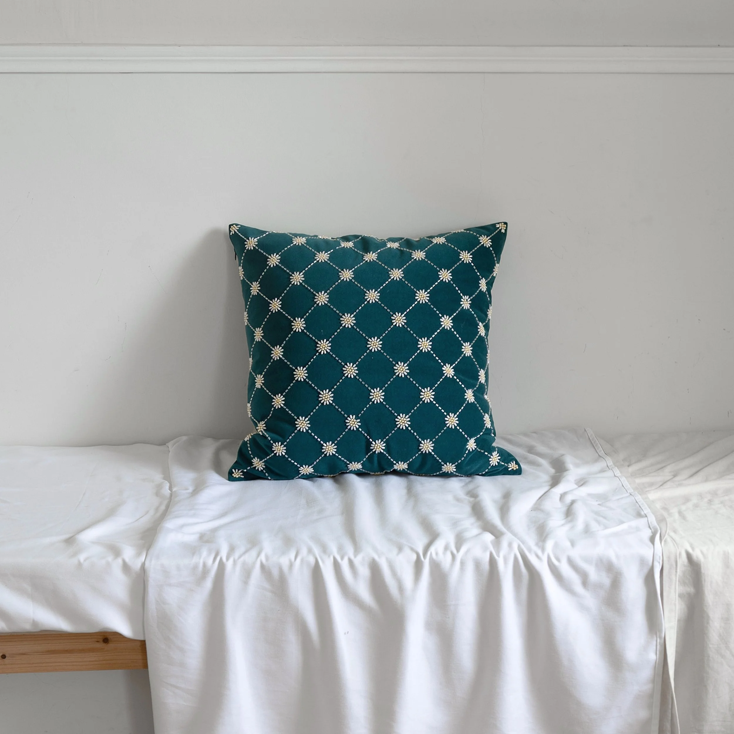 Teal velvet Daisy throw pillow with beading embroidery styled on a bed with white bedding in a minimalist bedroom.