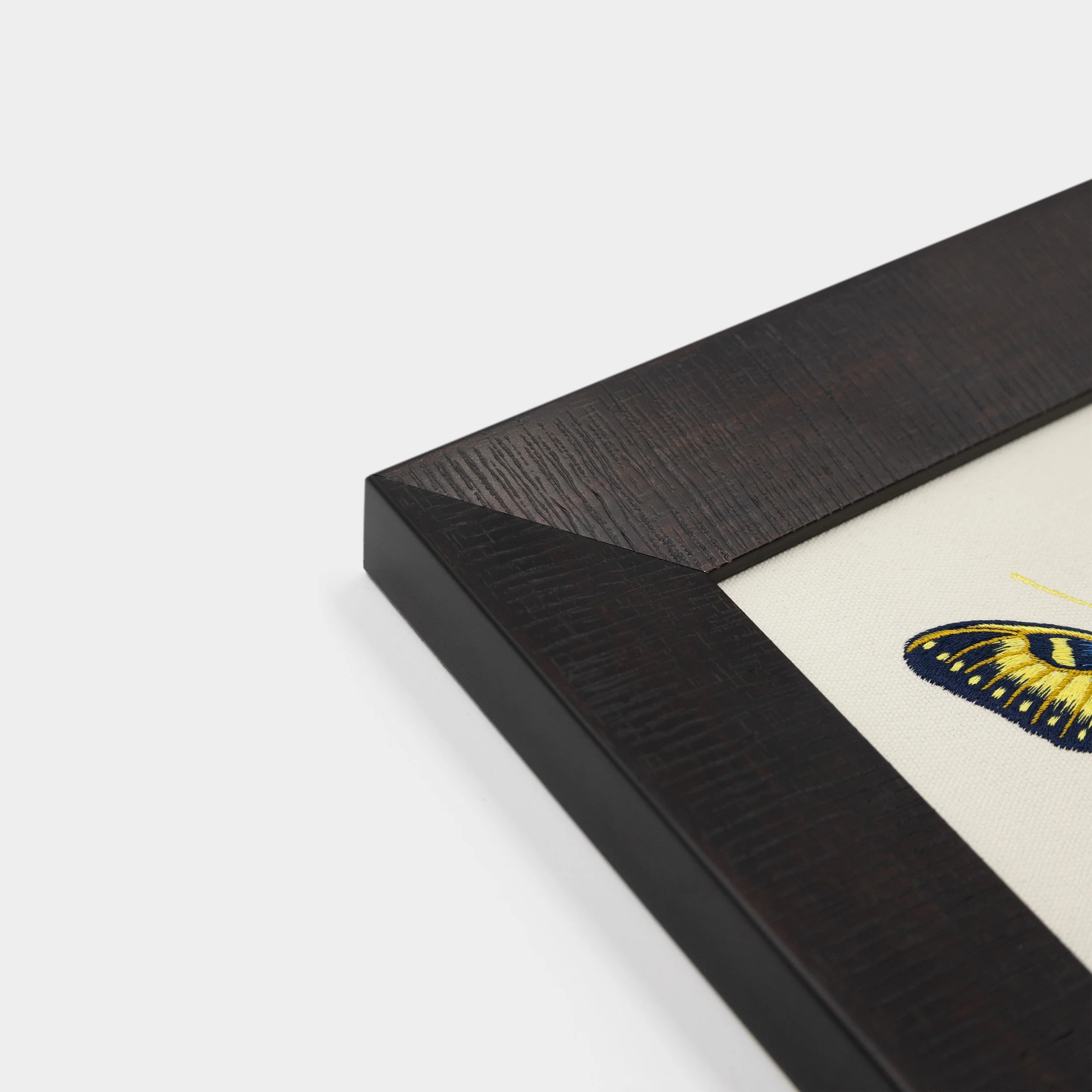 Corner detail of the dark solid wood frame surrounding the Luminara butterfly embroidered artwork.