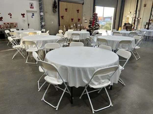 Round tables with white linens and folding chairs arranged for a large indoor event