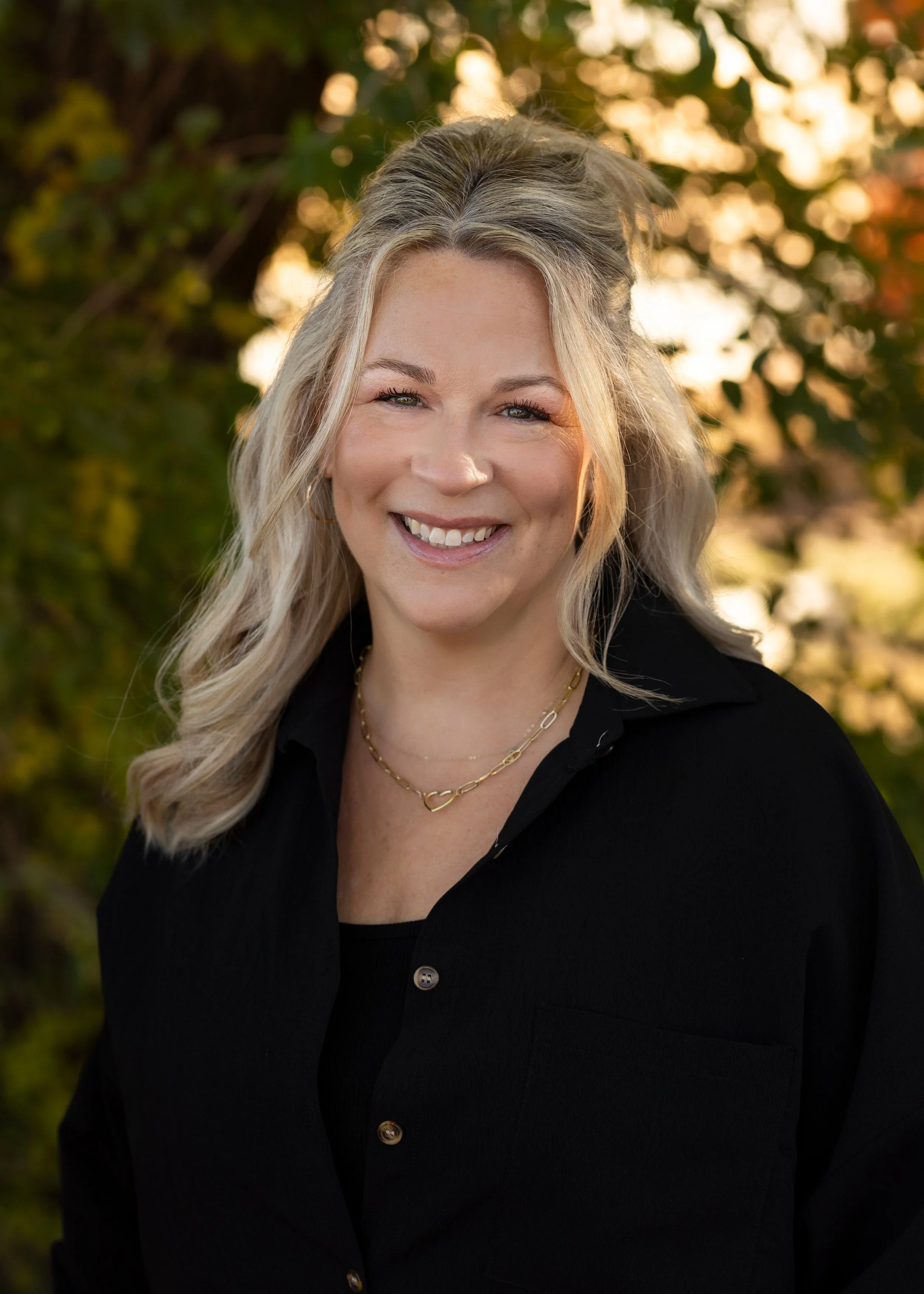 A Blooms staff member with blonde hair wearing a black top standing outdoors during sunset with trees in the background.