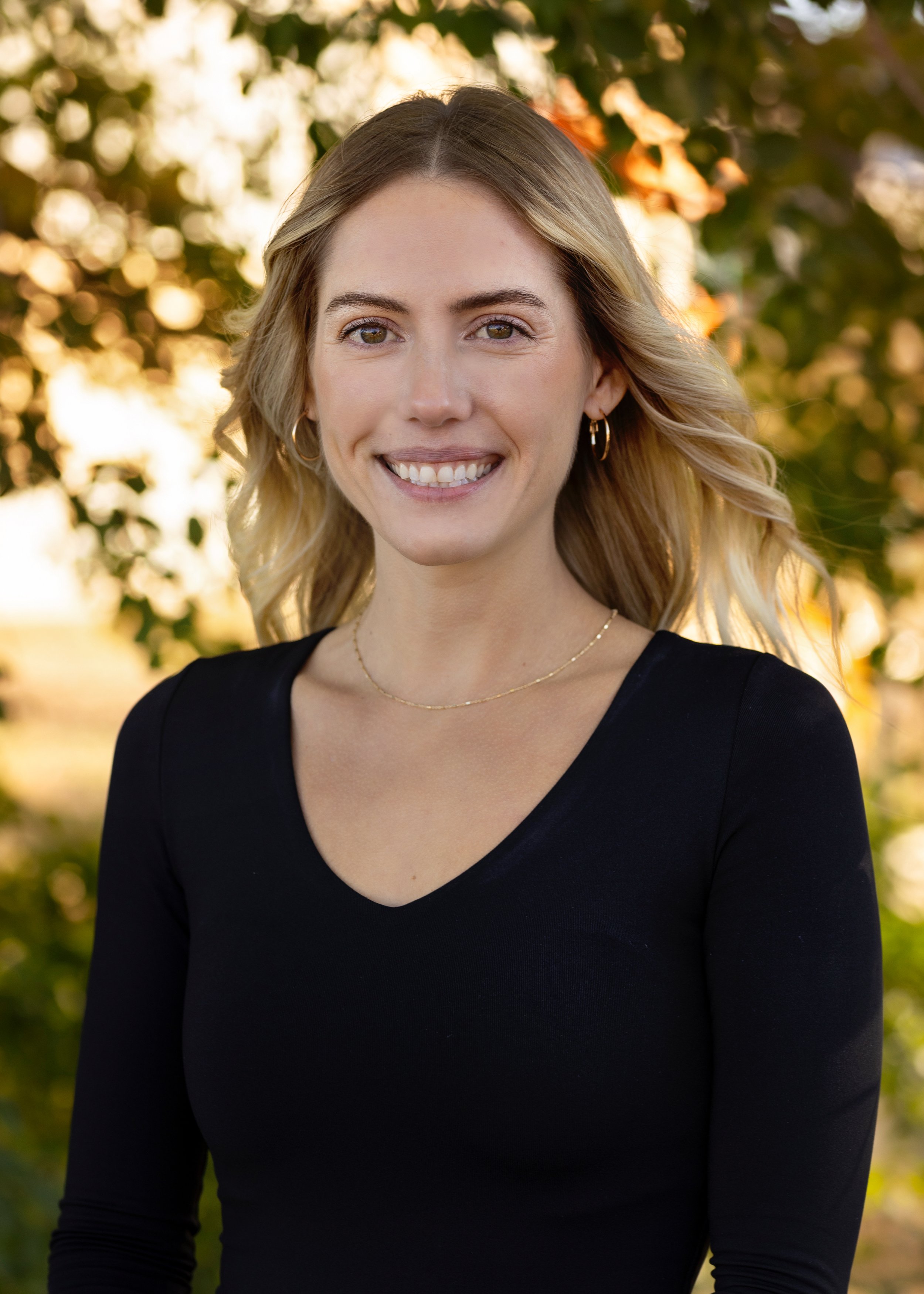A Blooms staff member with blonde, wavy hair, smiling, wearing a black top and gold jewelry, standing outdoors with sunlight filtering through trees in the background.