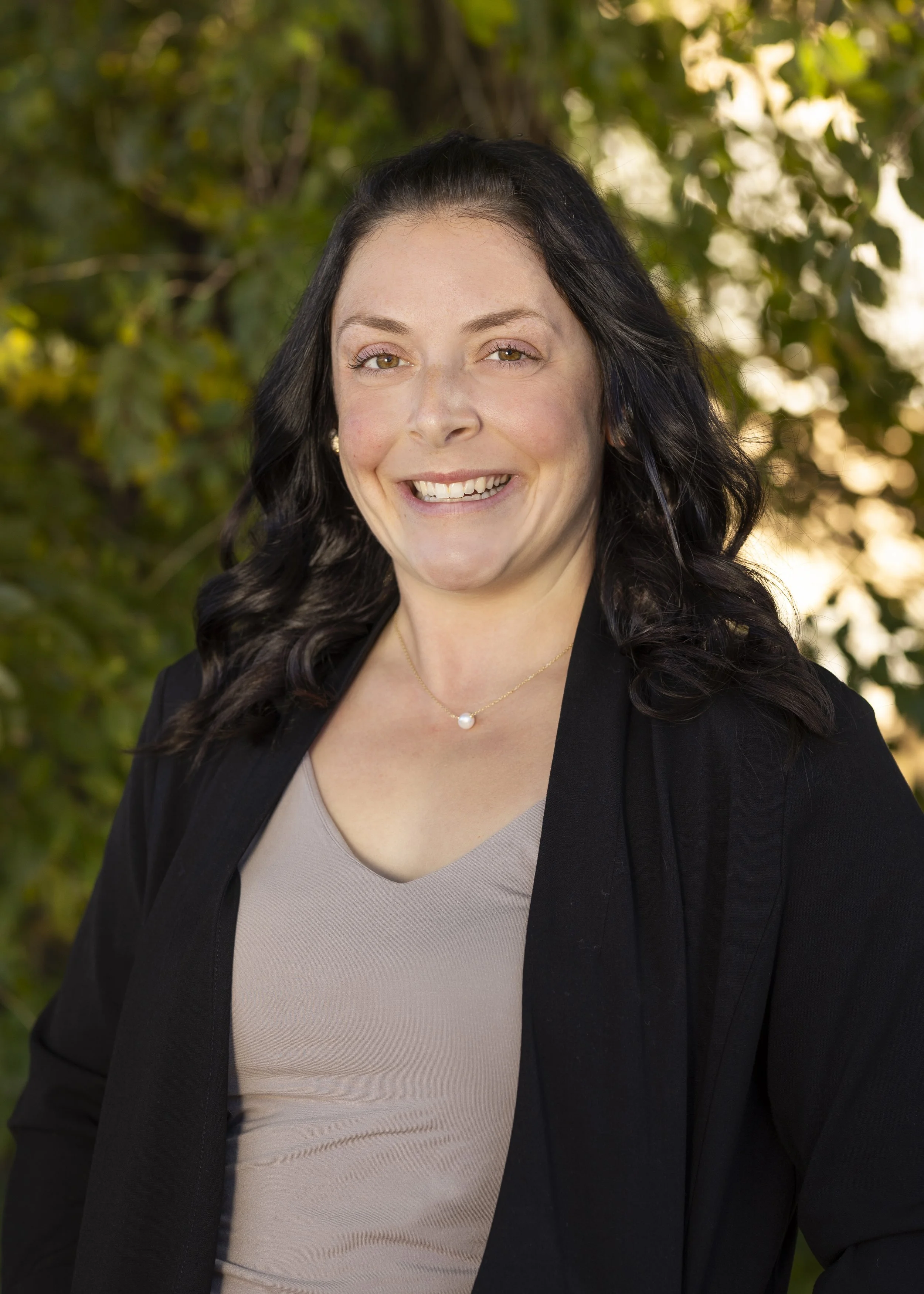 A Blooms staff member with black wavy hair, wearing a black blazer and a light beige top, smiling outdoors with trees and sunlight in the background.