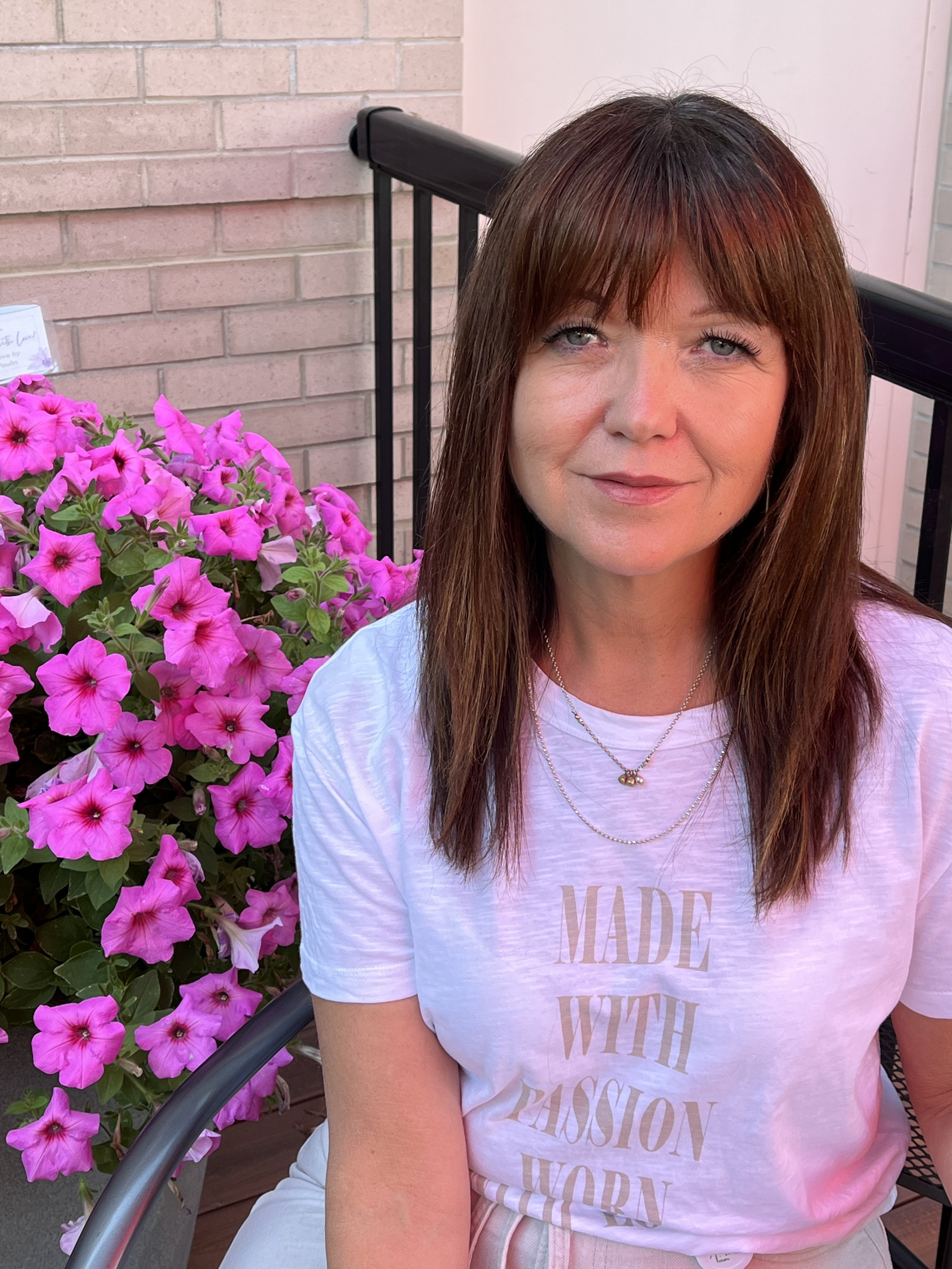 A Blooms Board member with brown hair and blue eyes sitting on a patio next to pink petunias, wearing a white t-shirt with gold text and layered necklaces.