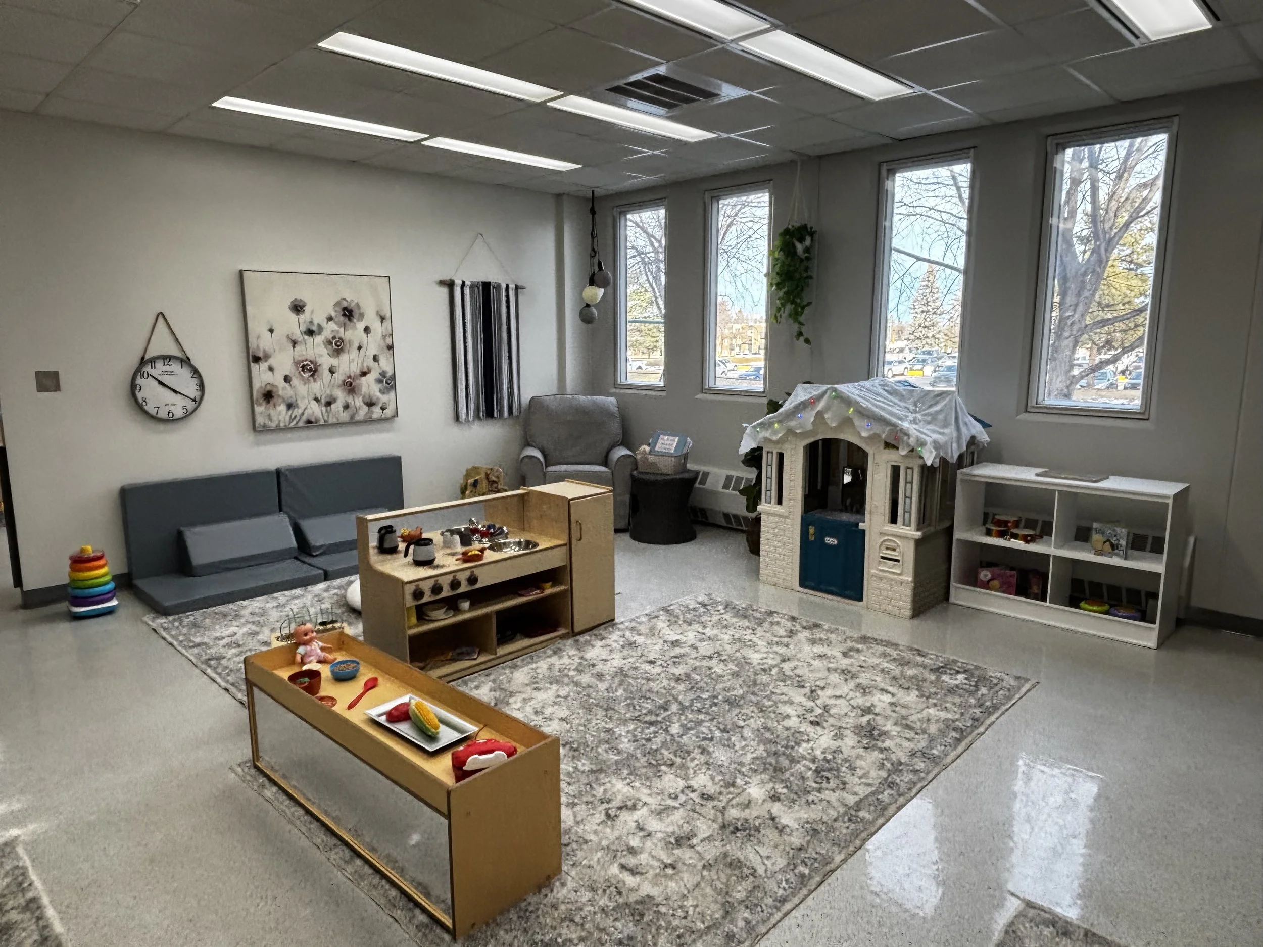 An early learning centre with a gray sitting area, a gray armchair, hanging plants, large windows, toys on a carpet, and a small wooden play kitchen.