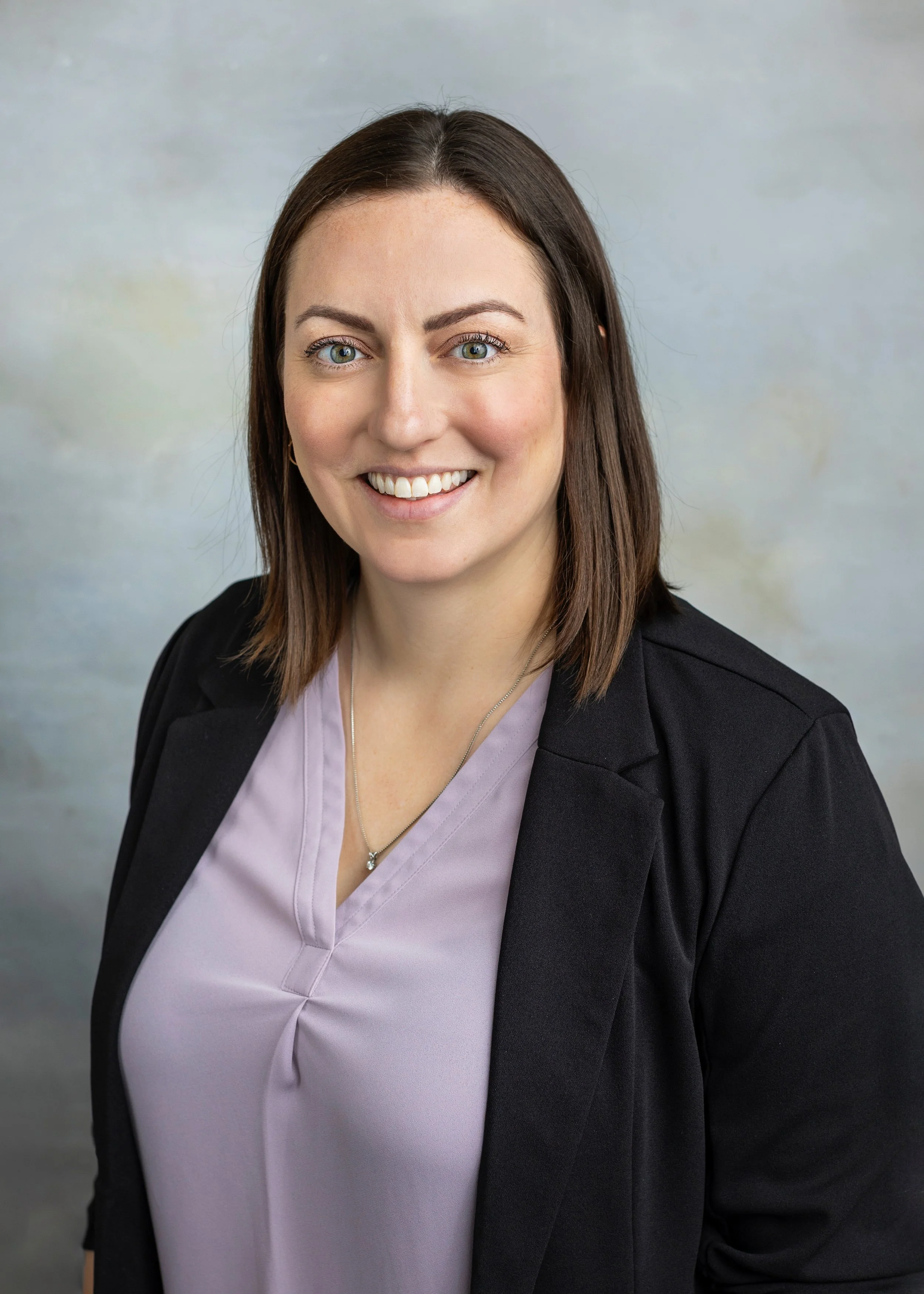 A Blooms Board member with shoulder-length brown hair, blue eyes, wearing a light purple blouse and a black blazer, smiling against a plain light background.