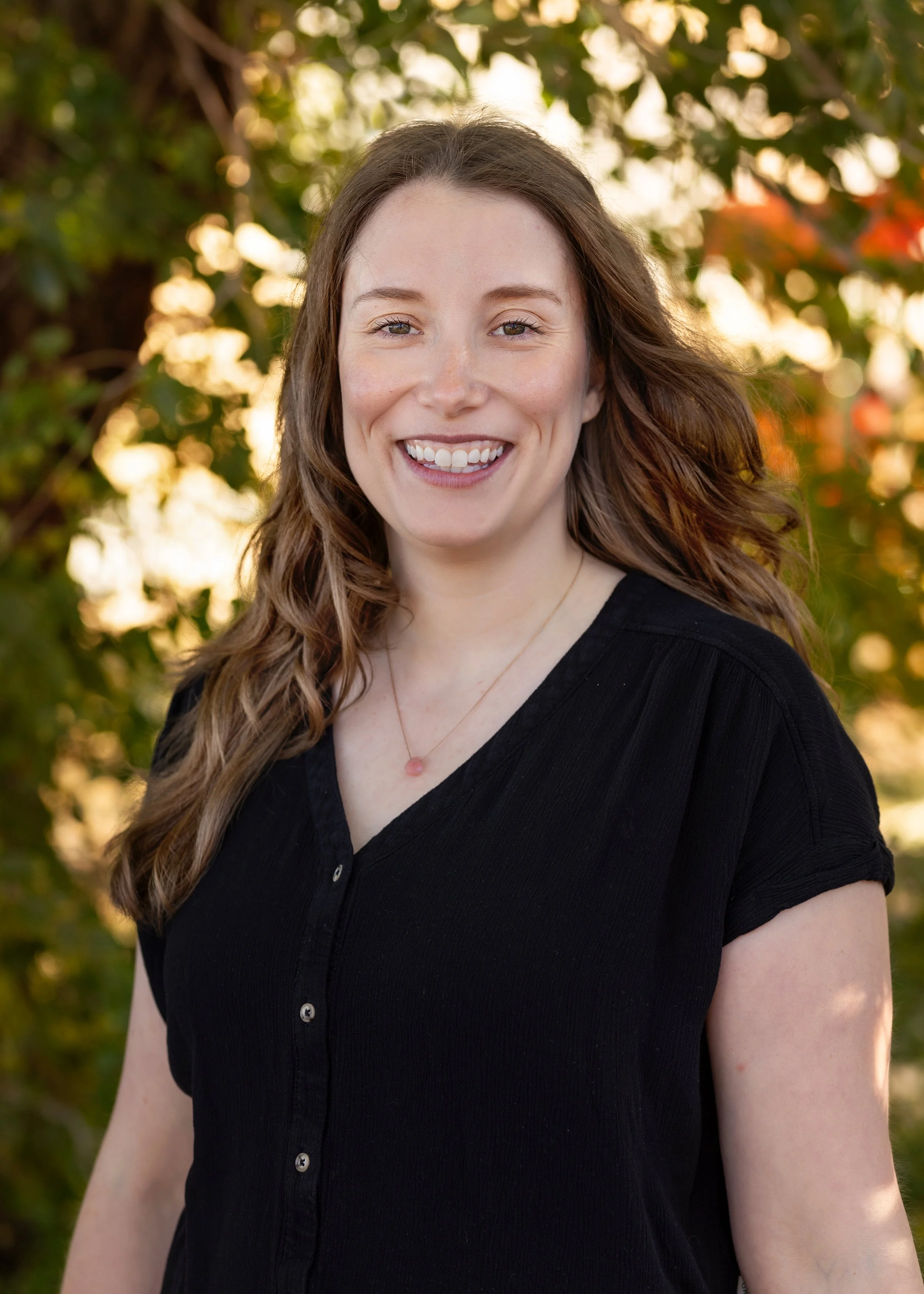 A Blooms staff member with long wavy brown hair smiling outdoors, wearing a black button-up shirt and a delicate necklace, with sunlight filtering through green foliage in the background.