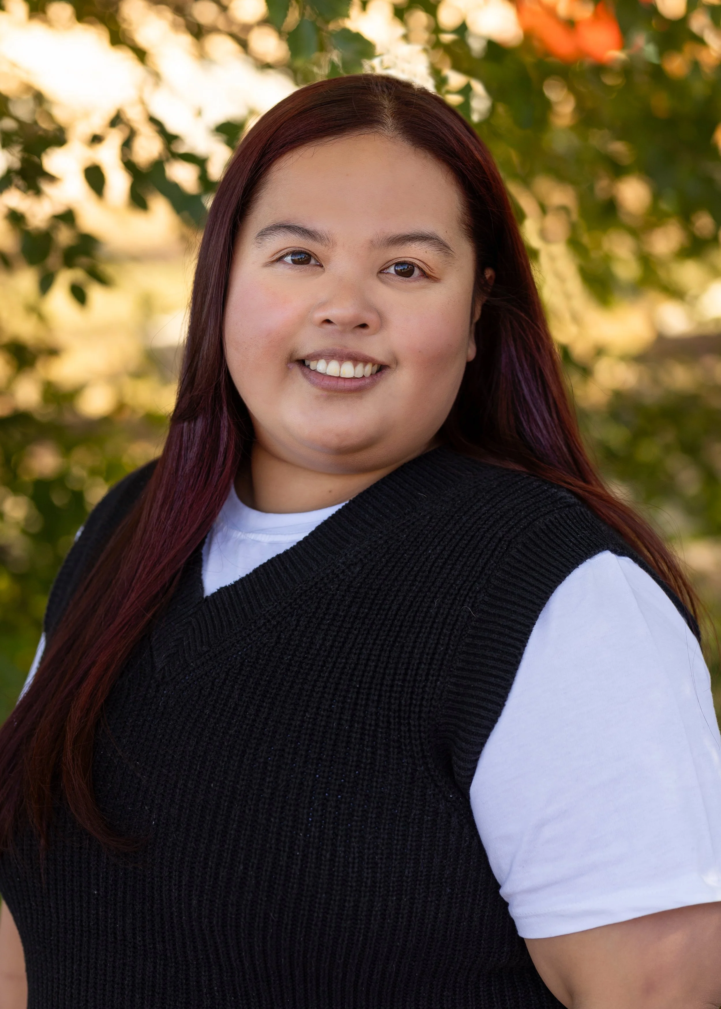 A Blooms staff member with long dark red hair smiling outdoors with blurred green foliage in the background.