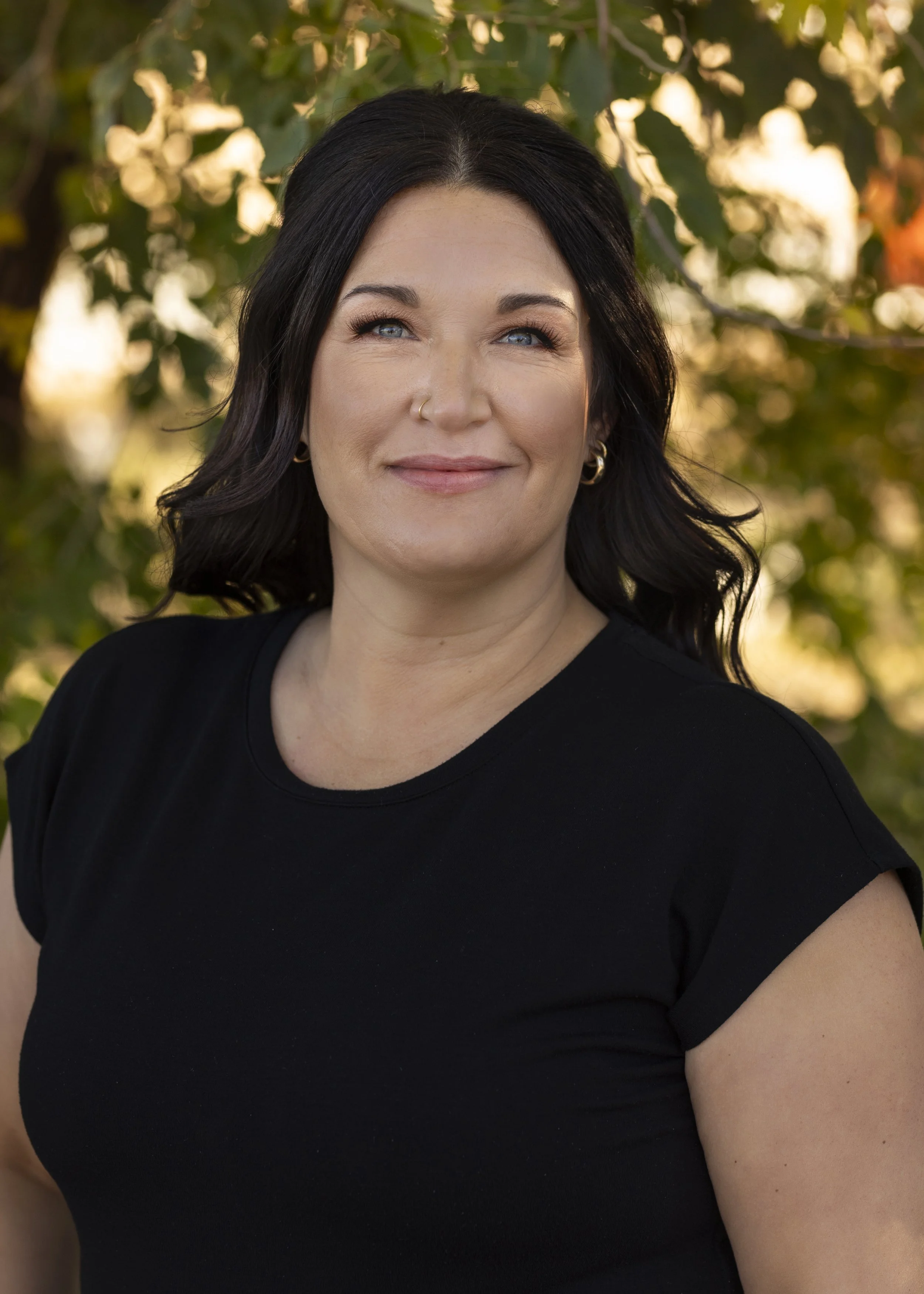 A Blooms staff member with black hair, blue eyes, and light skin, smiling outdoors with trees in the background.