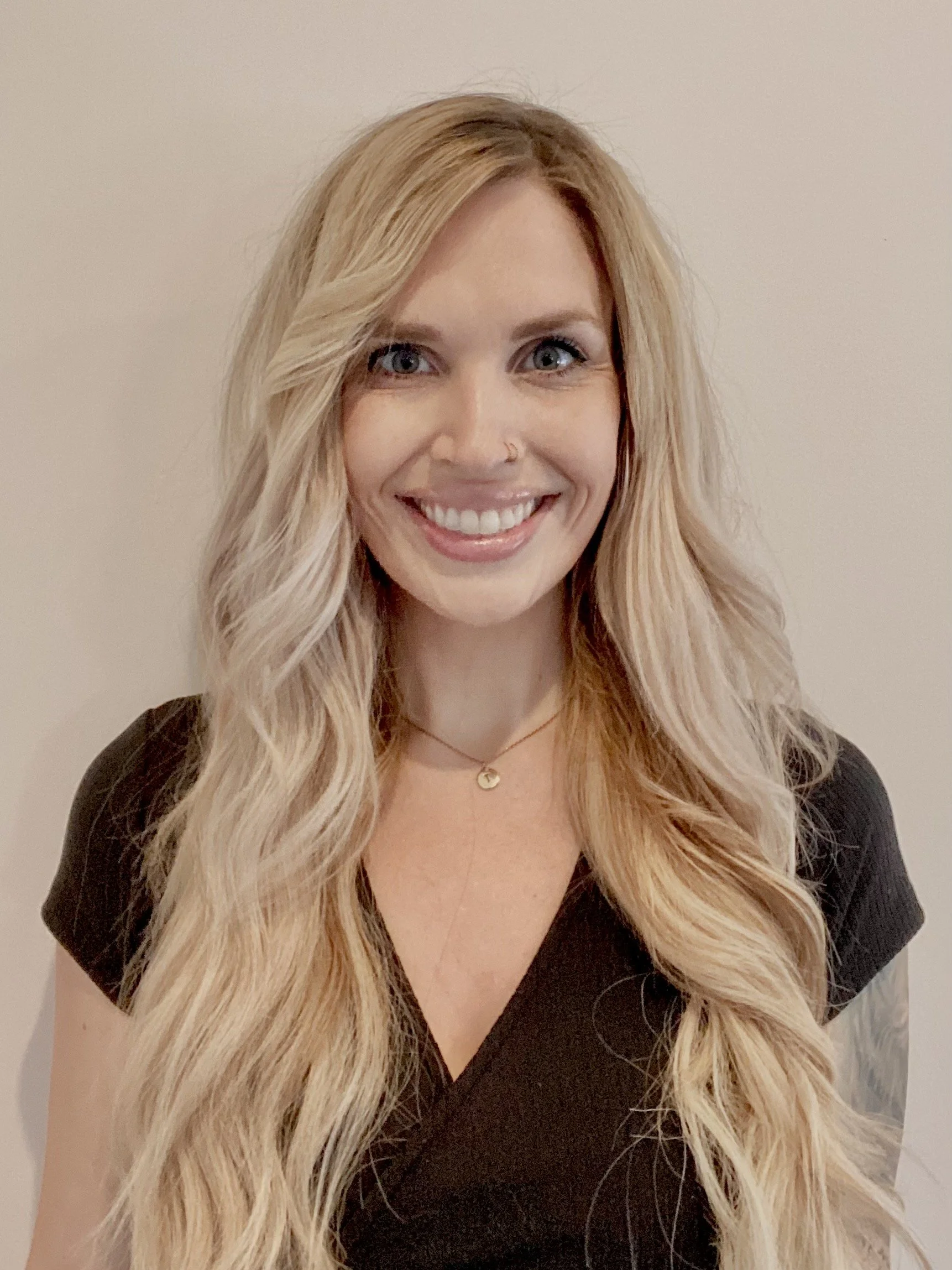 A Blooms Board member with long, wavy blonde hair, blue eyes, and a nose piercing, smiling and wearing a black top with a small necklace, standing against a plain light-colored wall.