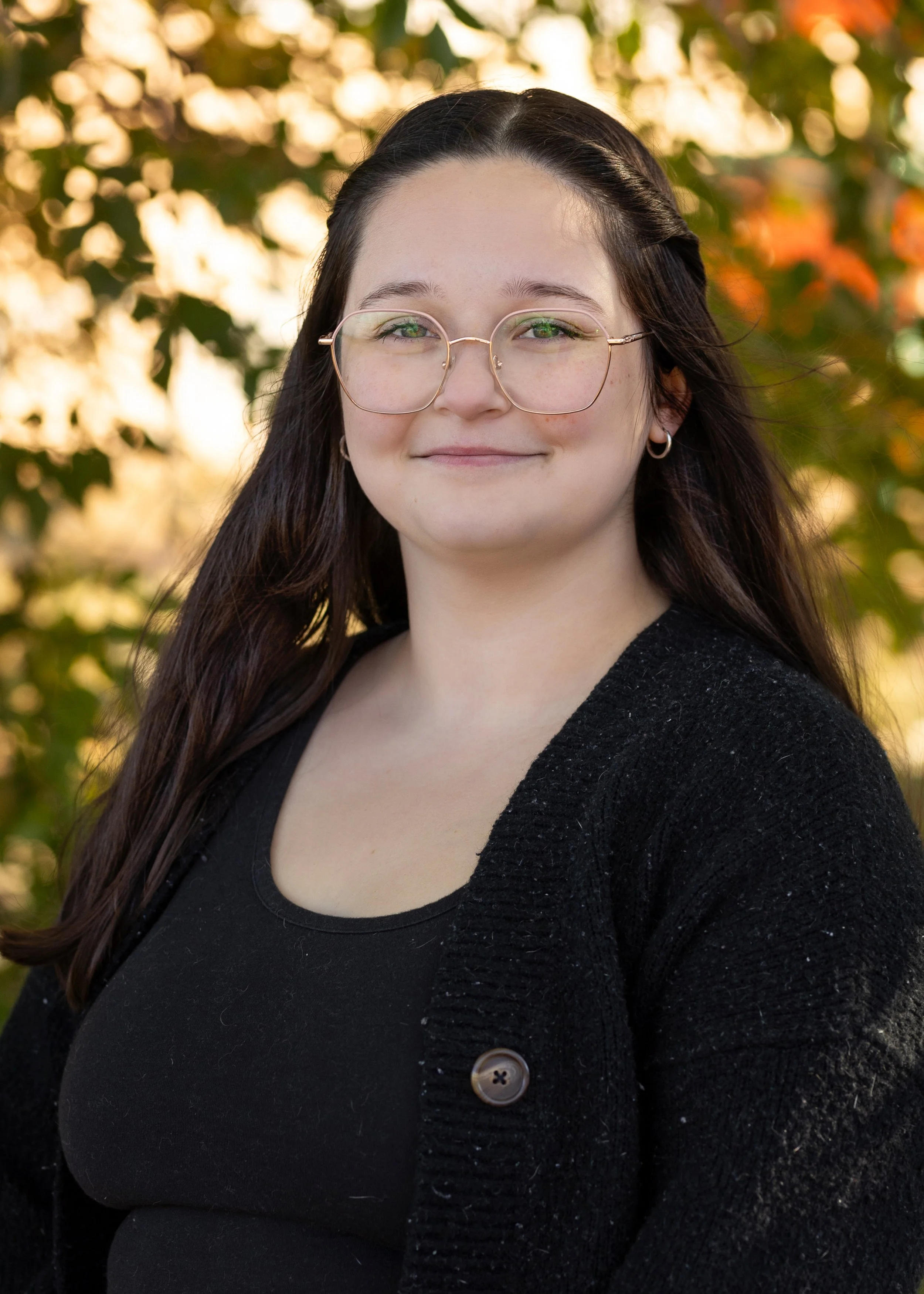A Blooms staff member with long dark hair, wearing glasses, hoop earrings, a black top, and a black cardigan, standing outdoors with autumn-colored leaves in the background.