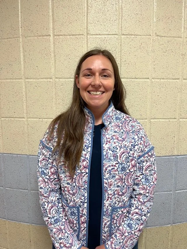 A Blooms Board member with long brown hair smiling, standing in front of a beige and gray cinder block wall, wearing a patterned jacket with blue and pink designs.