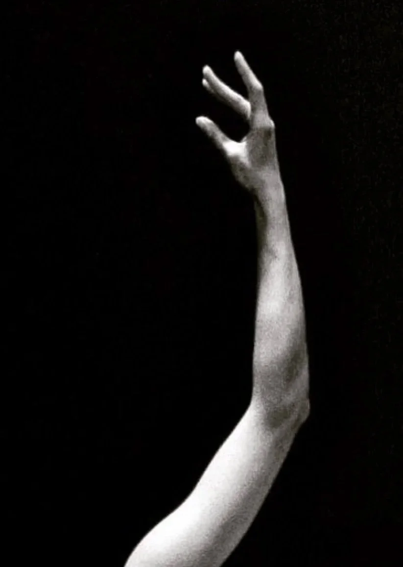 A black and white photo of a dancer's arm and hand reaching upward against a dark background